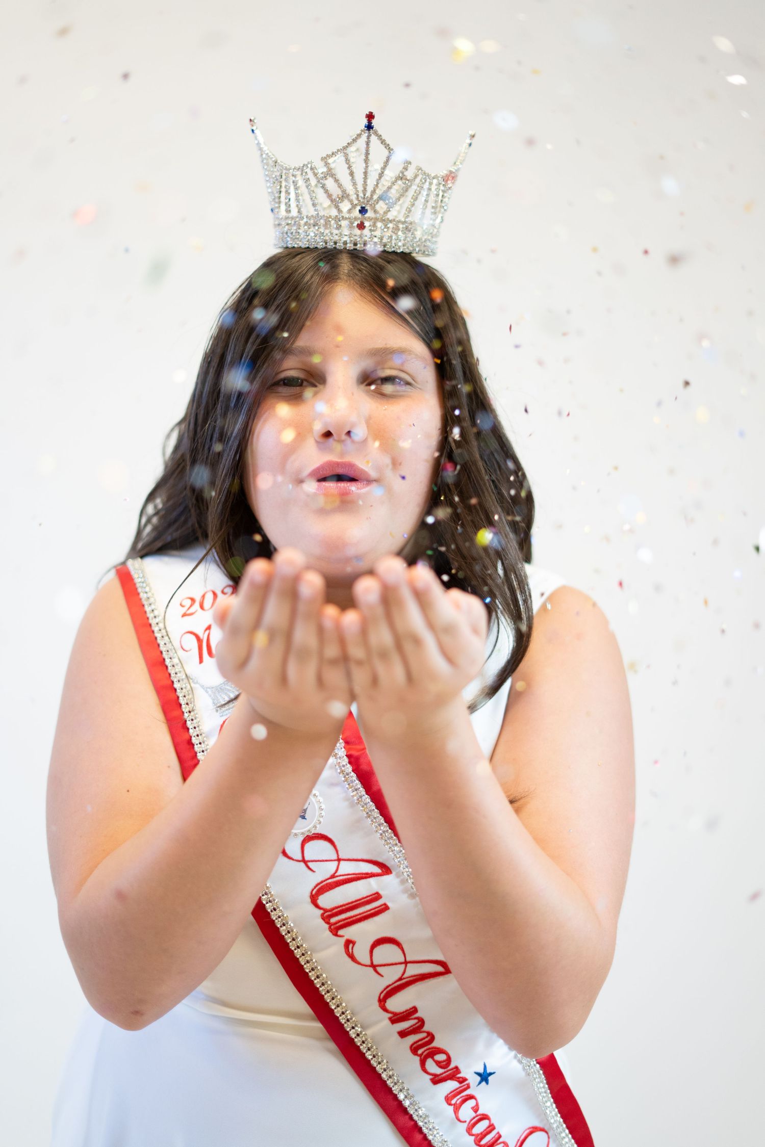 Pageant winner blowing glitter and wearing crown against white background.