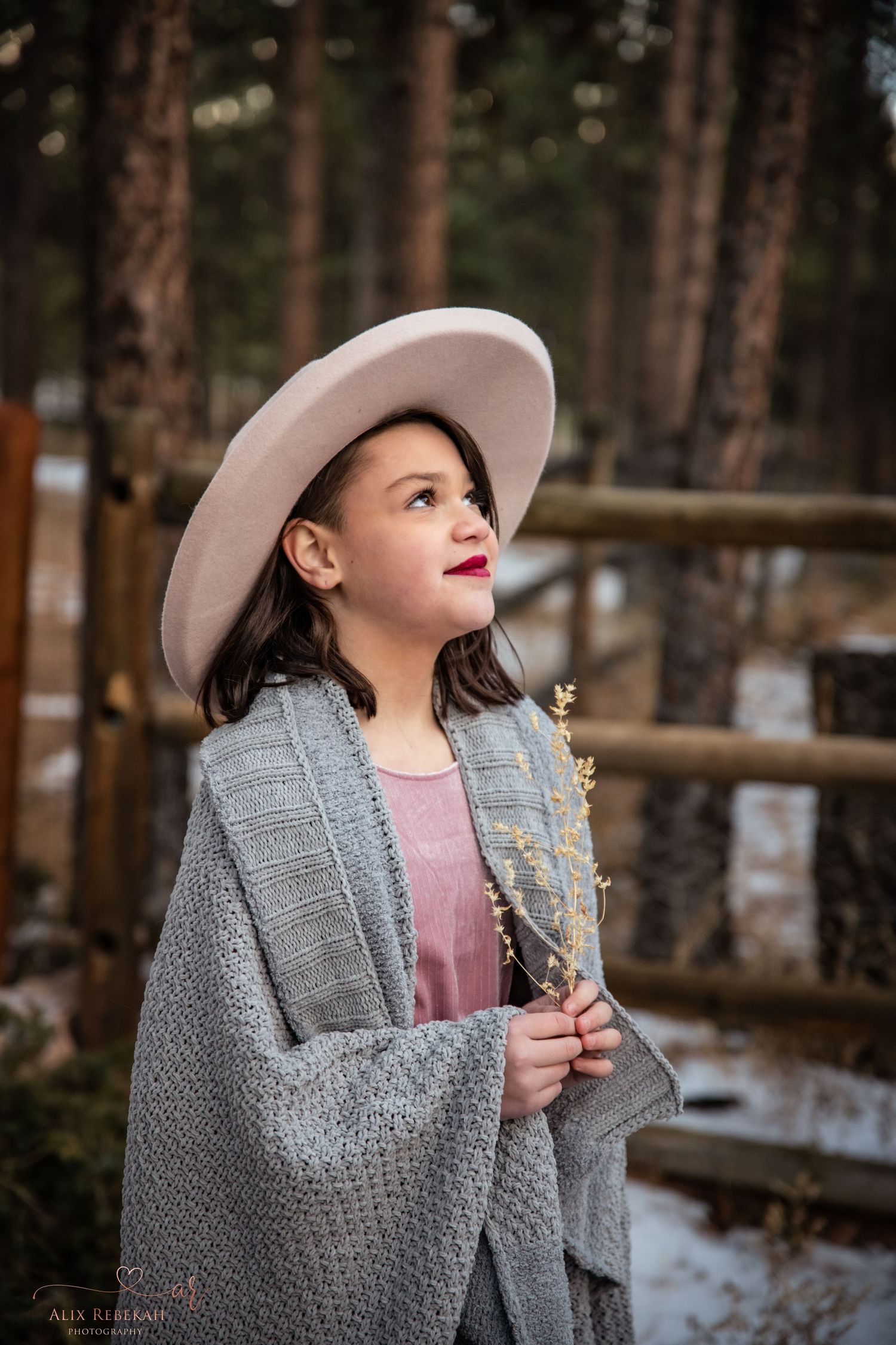 Person wearing a gray sweater and beige hat standing near wooden fence in forest with light snow on ground.
