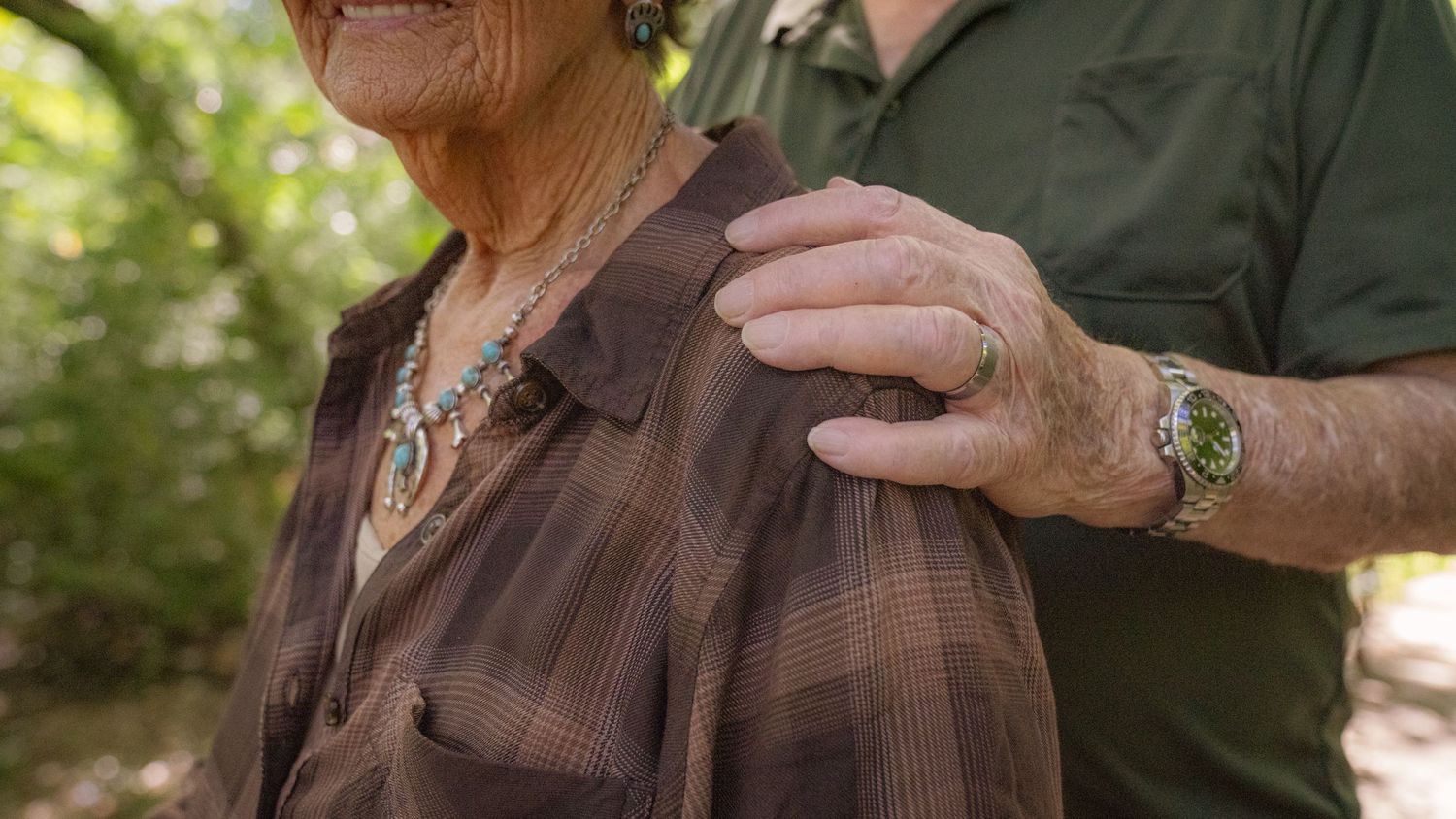 A close-up shot shows gentle hands resting on shoulders in a natural outdoor setting.