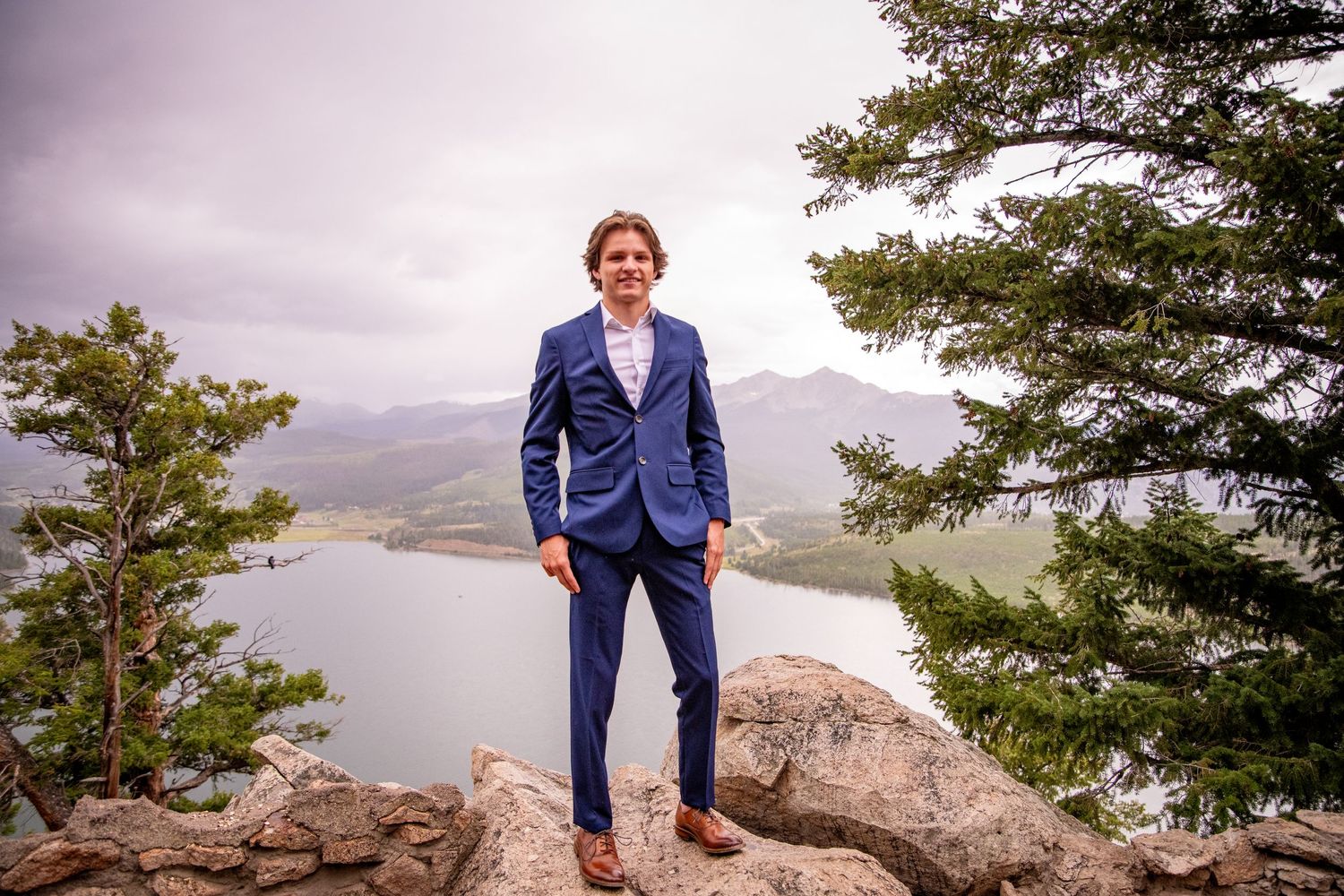 Navy suited figure standing on rocky overlook with lake and mountains in background.