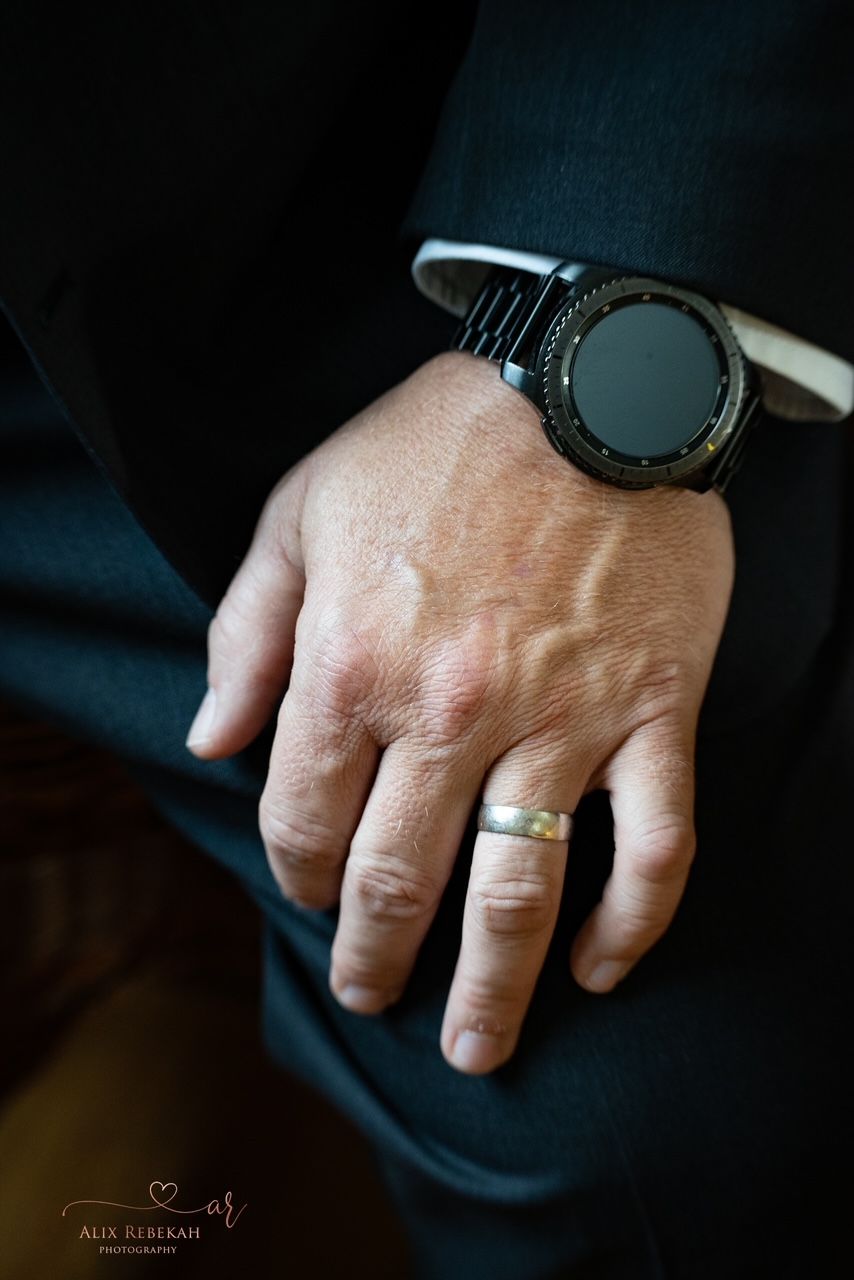 A hand wearing a black digital watch and silver wedding ring rests casually on dark fabric.