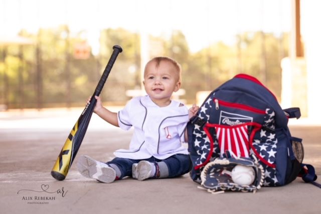 Baby baseball player sitting with bat and patriotic gear bag.