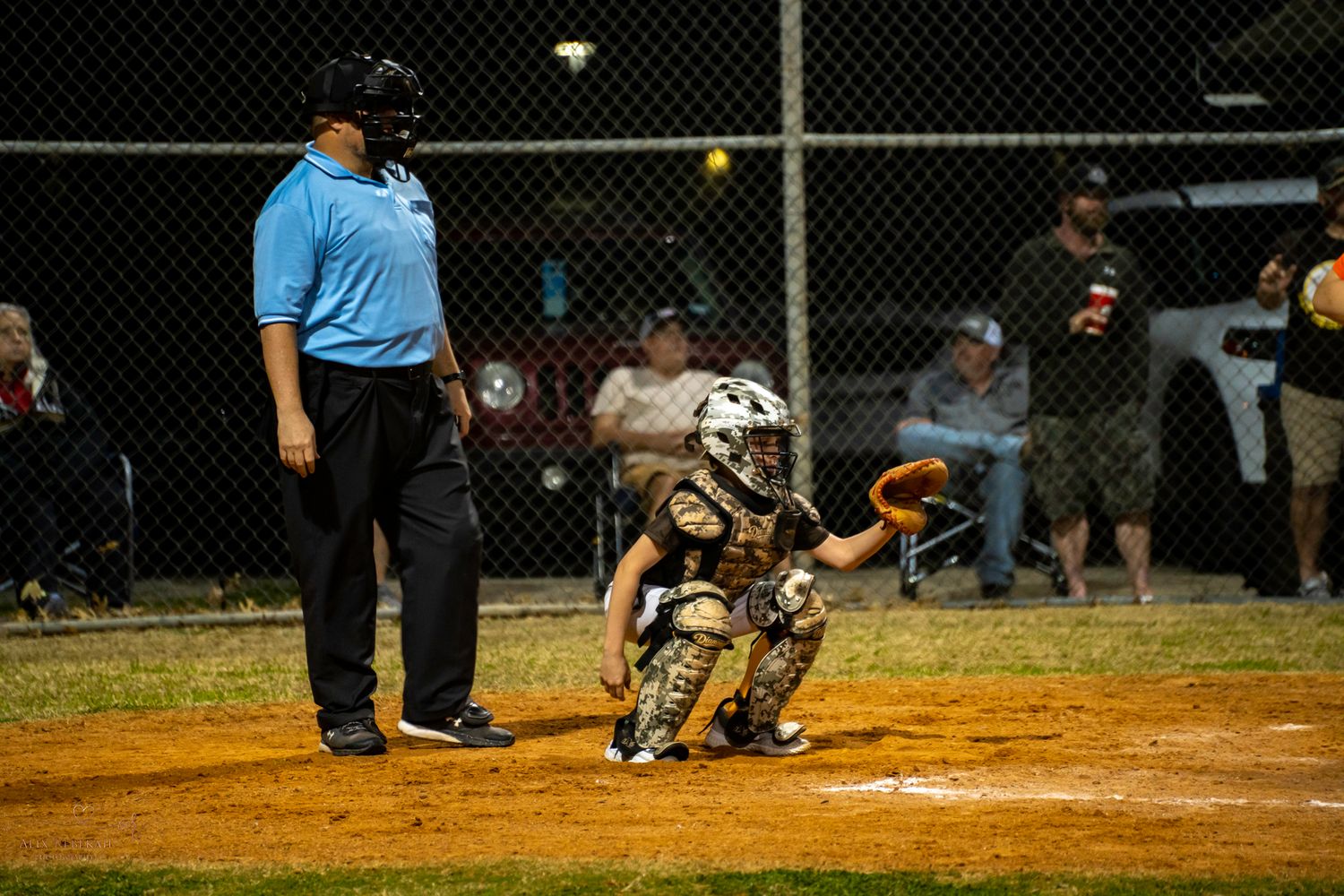 A baseball catcher crouches behind home plate at night as an umpire stands nearby during a game.