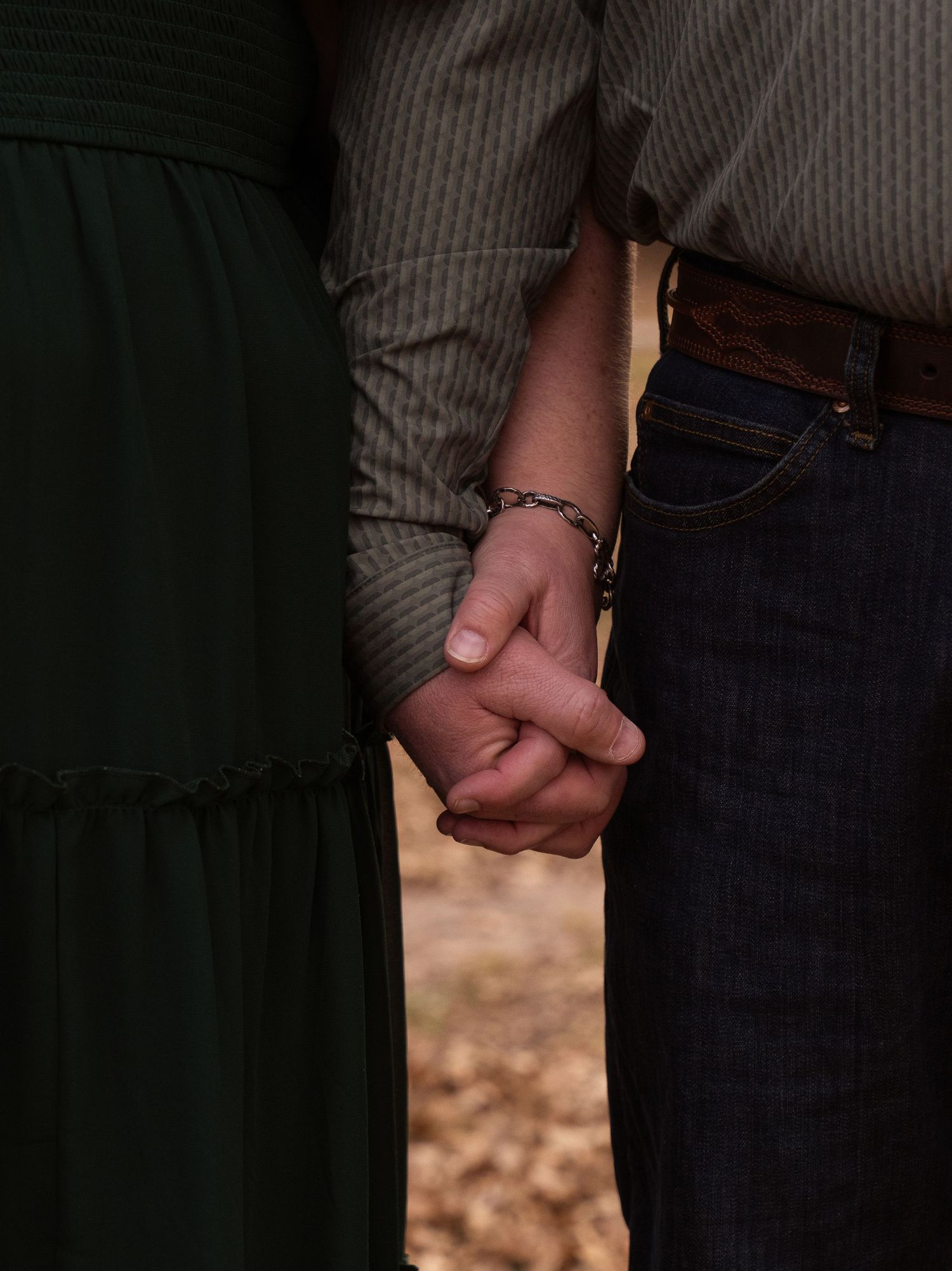 A couple holds hands tenderly, wearing green dress and jeans, with a bracelet visible on one wrist.