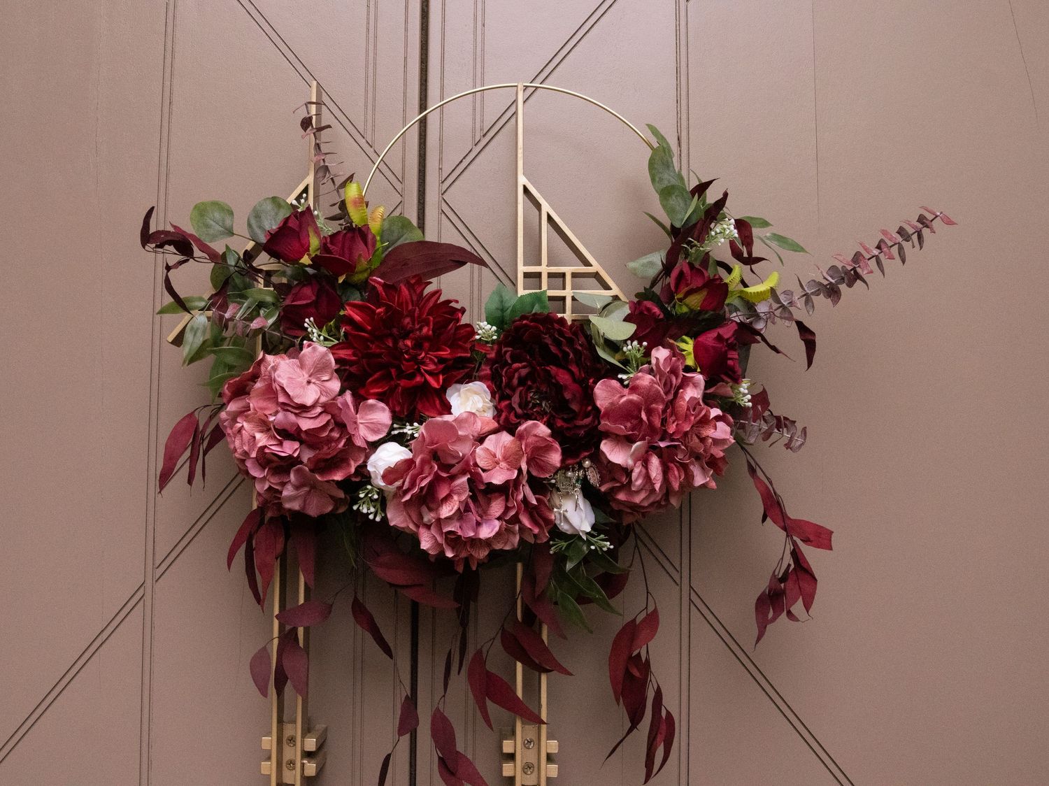 Elegant burgundy and pink floral wreath with hydrangeas and dahlias on a gold geometric frame against a beige wall.