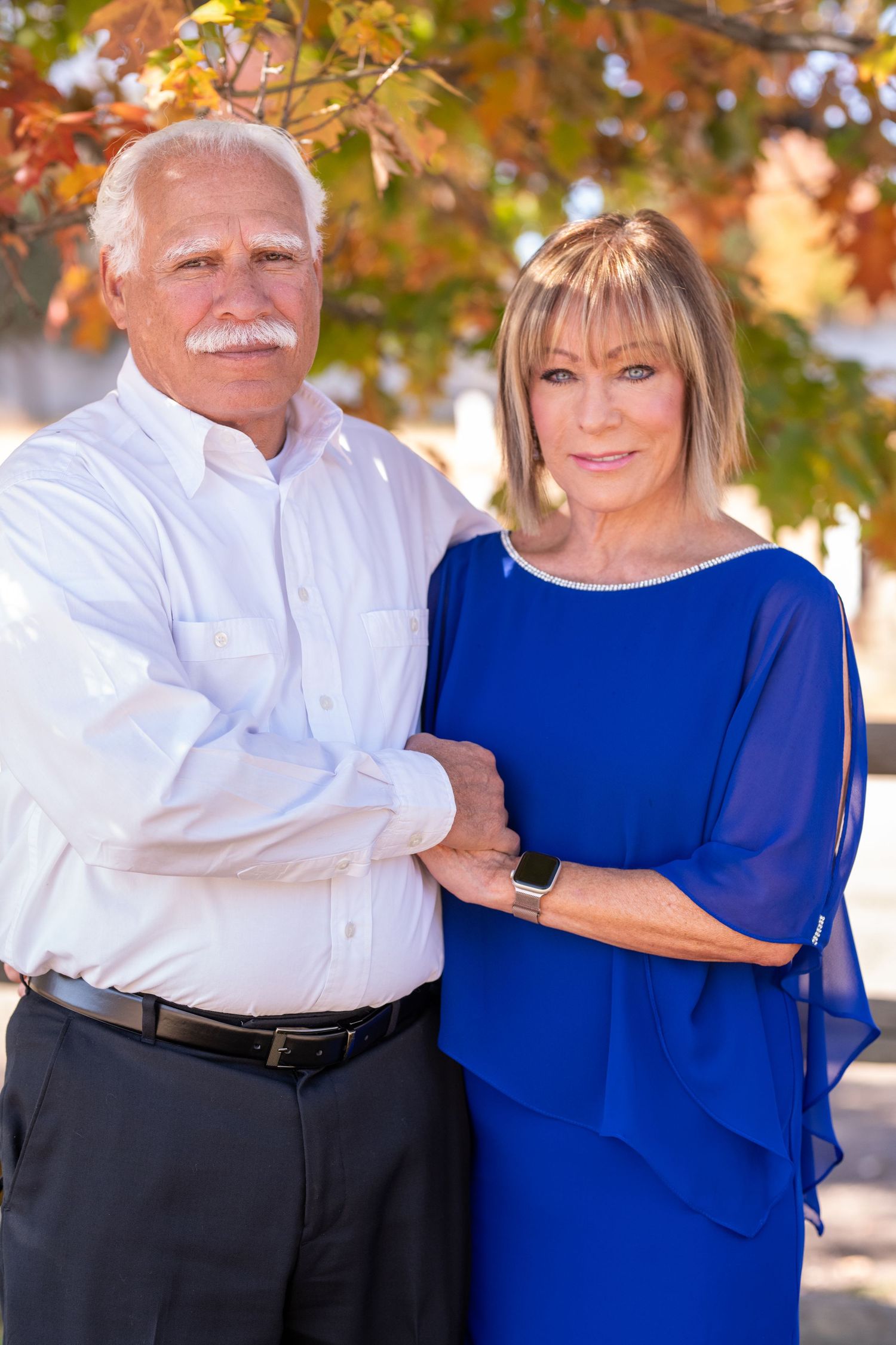 A couple poses outdoors in autumn, with the person in white and person in blue embracing against fall foliage.