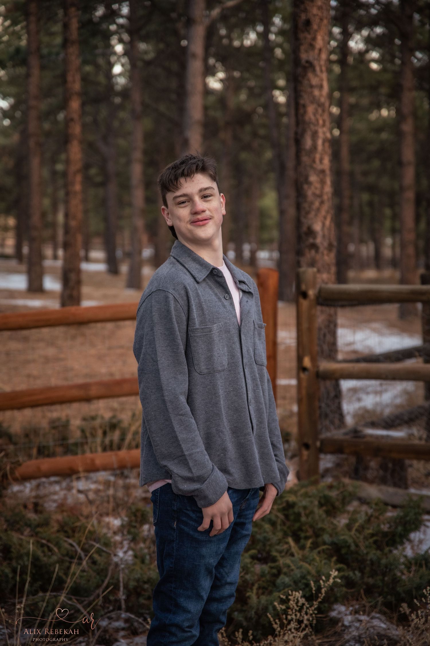 Person in gray jacket and jeans standing by wooden fence in snowy forest setting.