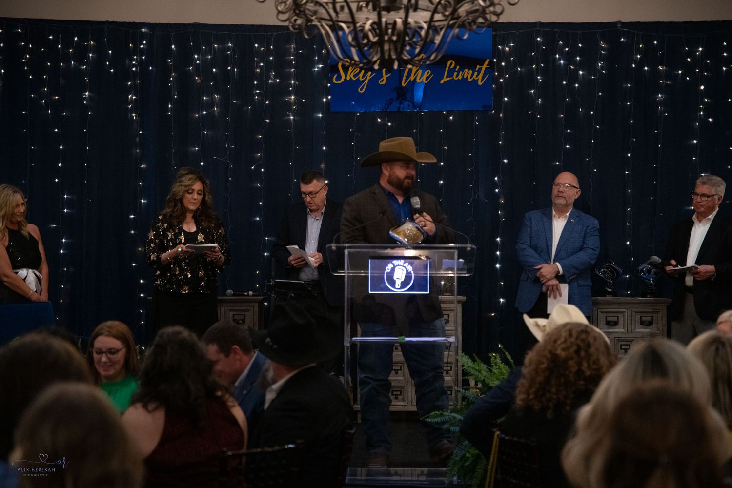 Speaker at podium addresses crowd during formal evening event with decorative lighting.