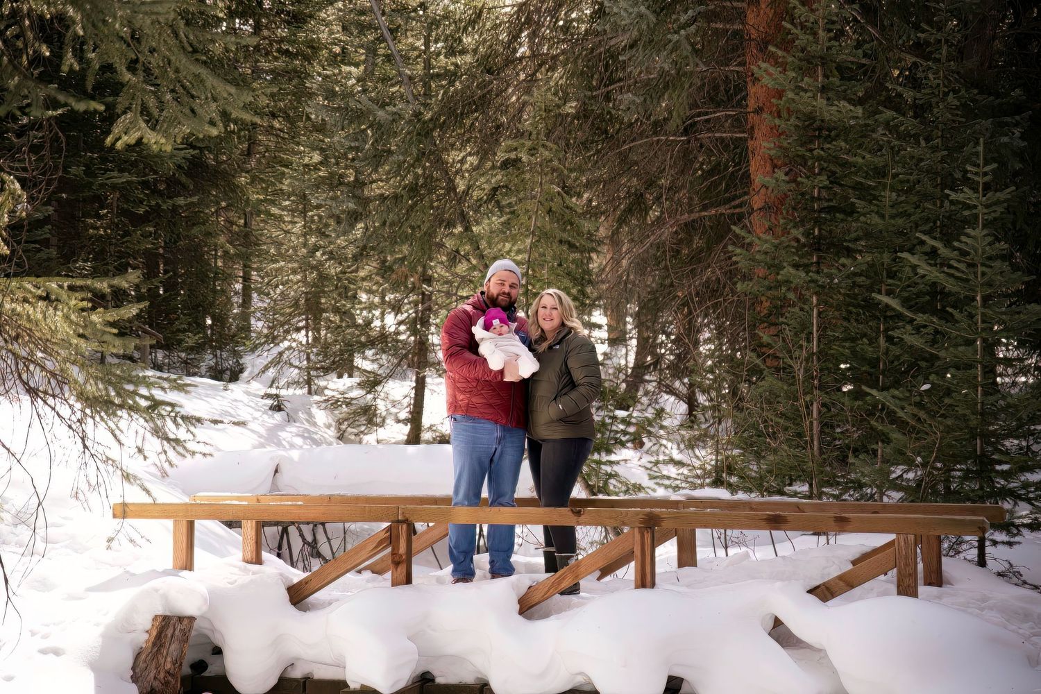 Two people stand together on a snowy wooden bridge in a winter forest setting.