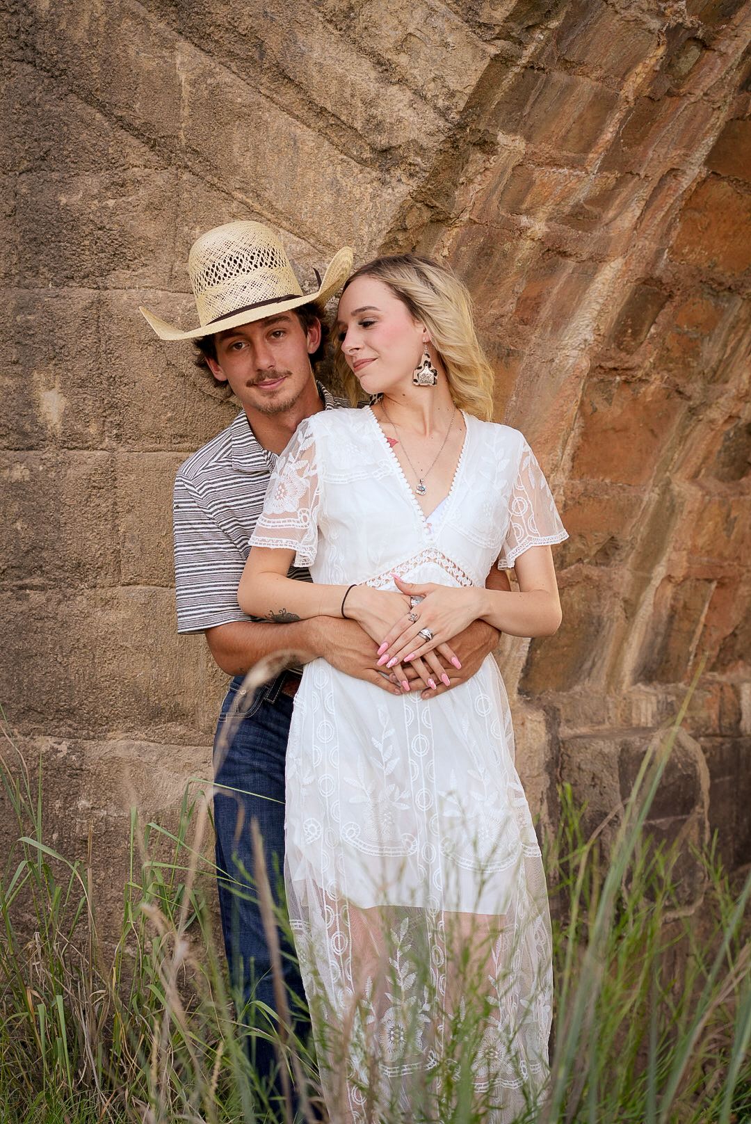 A couple embraces against a rustic stone wall with tall grass in the foreground during a romantic outdoor photoshoot.