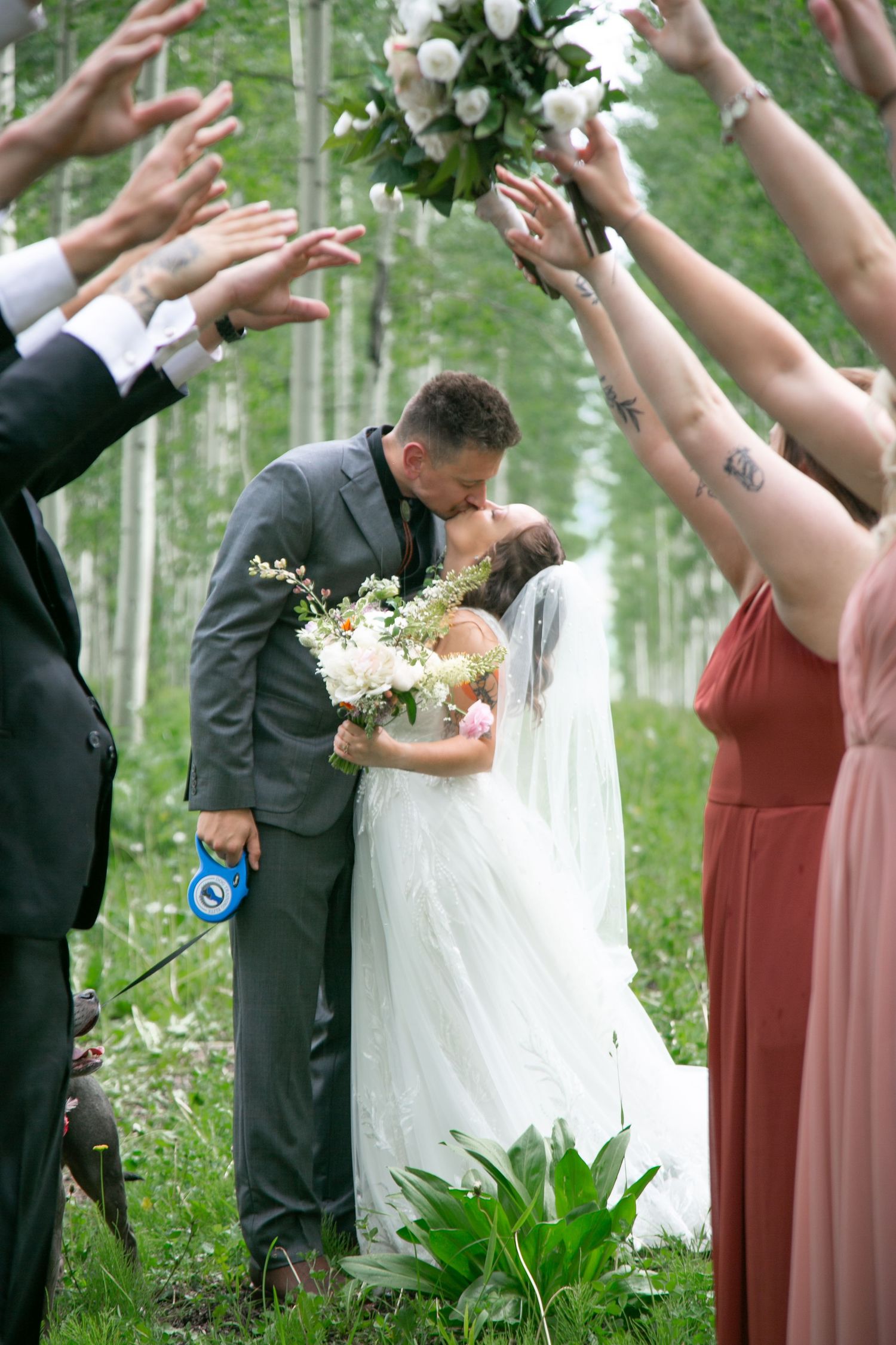 Wedding ceremony in forest with guests raising hands while couple kisses under floral arch.