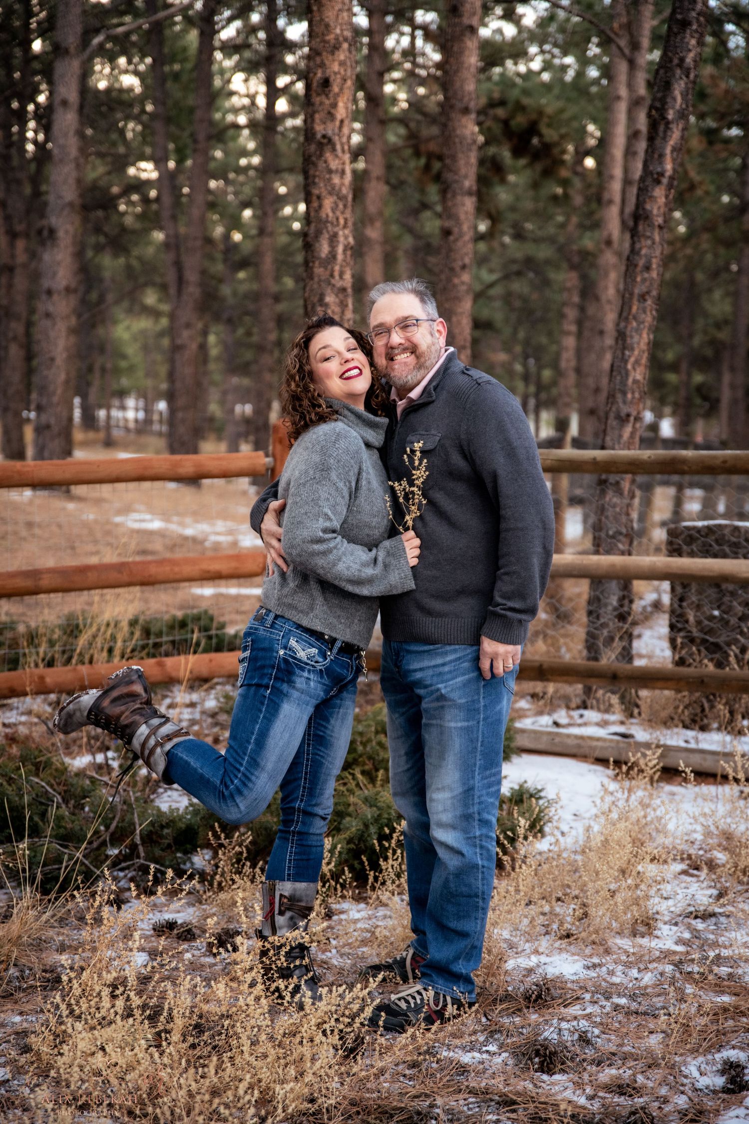Candid moment of couple laughing together by wooden fence in winter forest setting.
