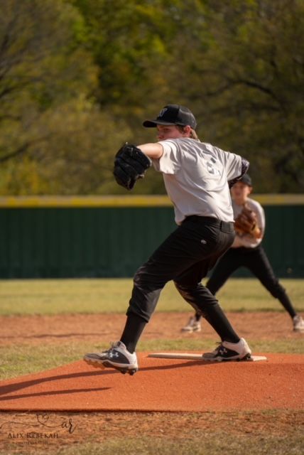 Baseball pitcher in white jersey and black pants winds up for a throw from the mound during a game on a sunny day.