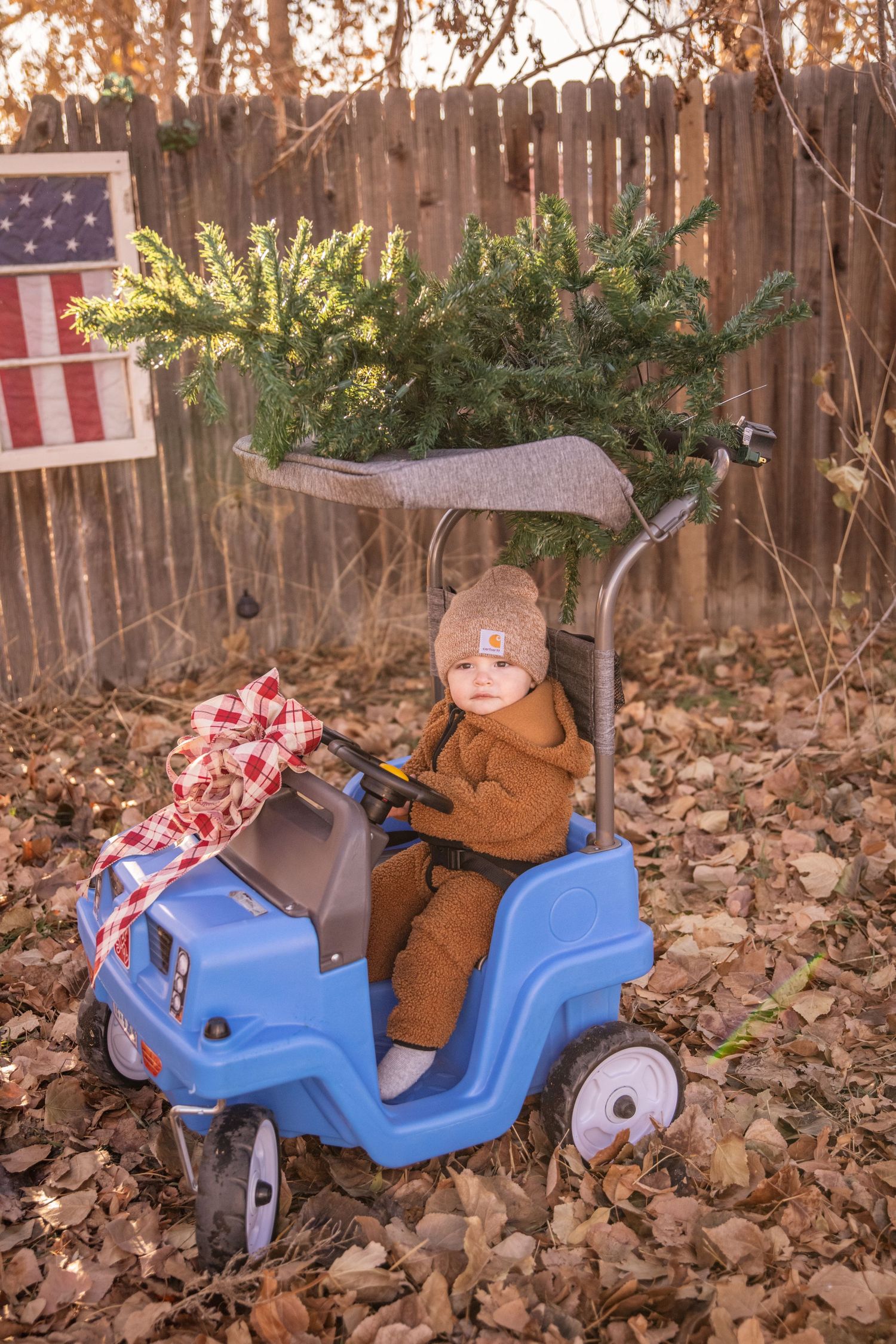 Small child in brown coat rides blue toy car with American flag in background.