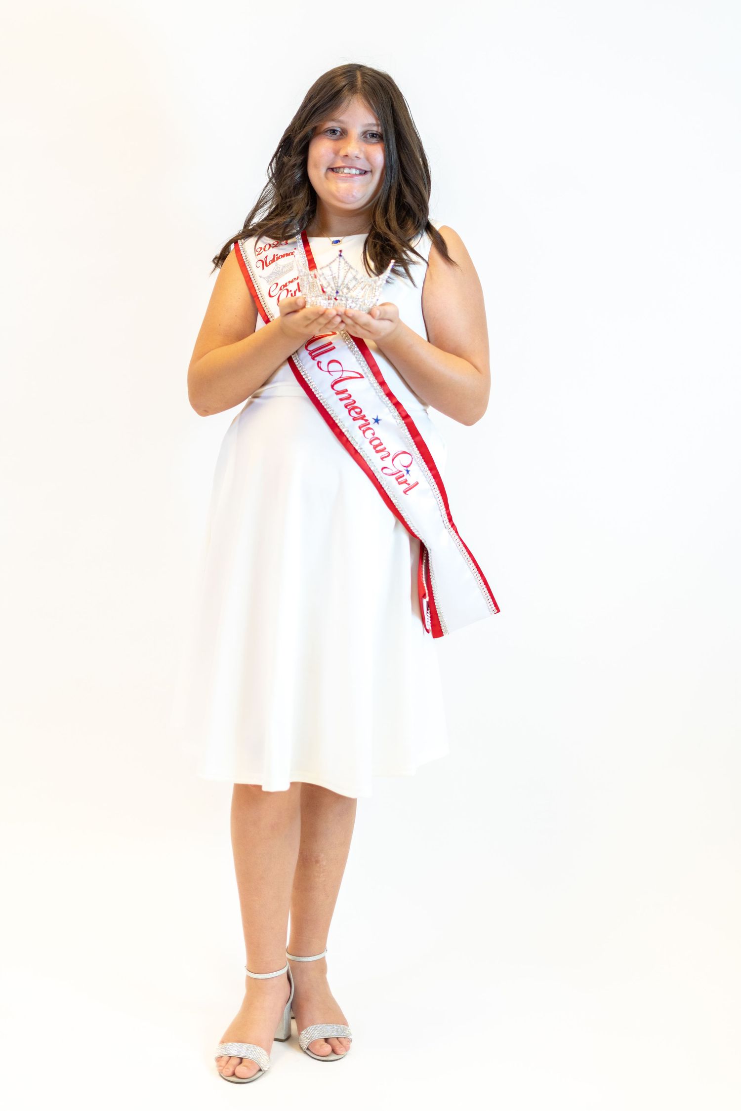 A contestant in a white dress poses with her pageant sash and crown against a white backdrop.
