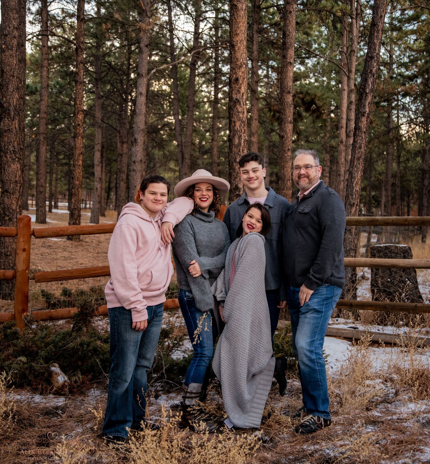Winter family portrait in pine forest with snow covered ground and wooden fence.