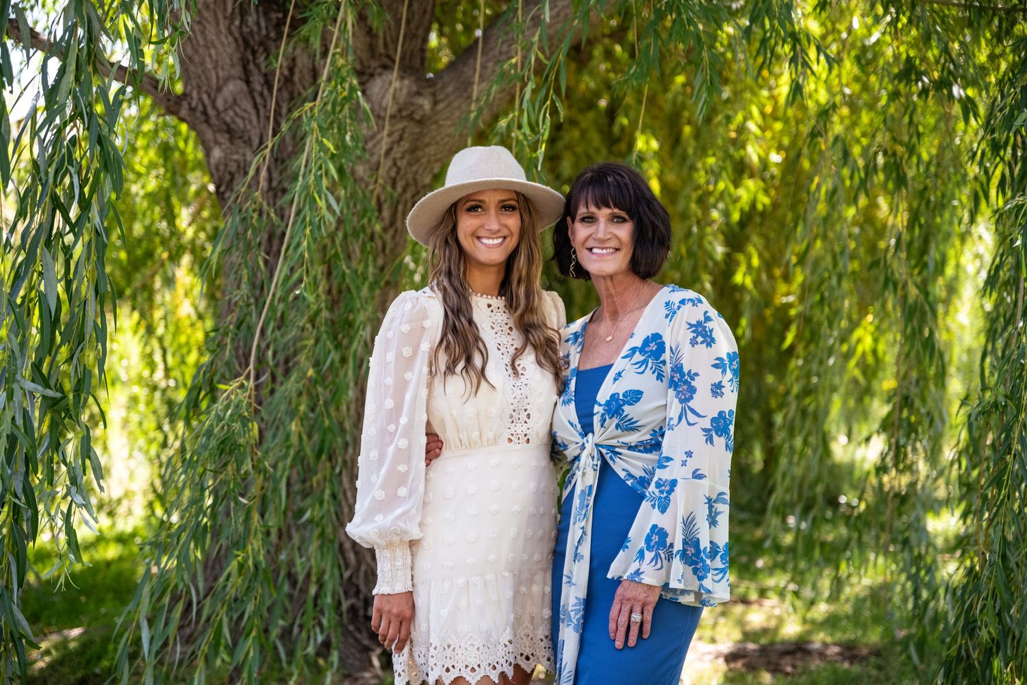 Two women in contrasting white and blue dresses pose under willow tree branches.