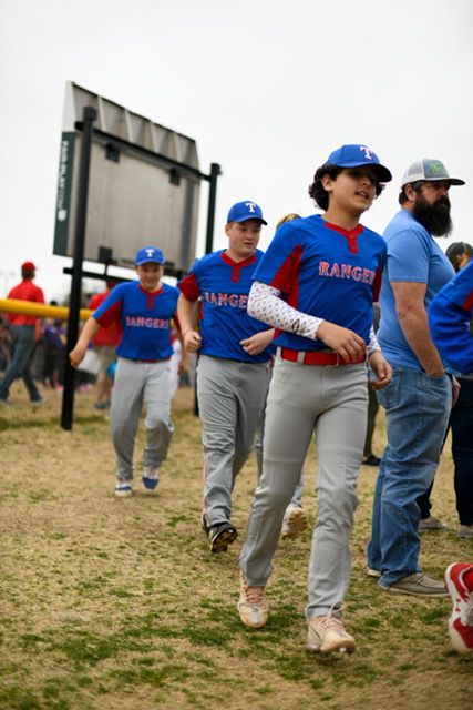 Baseball players in blue Rangers uniforms and grey pants walk across a grassy field during a team practice session.