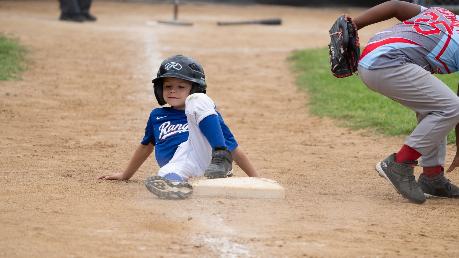 Youth baseball player in blue uniform slides into base during competitive game as opponent attempts tag play.