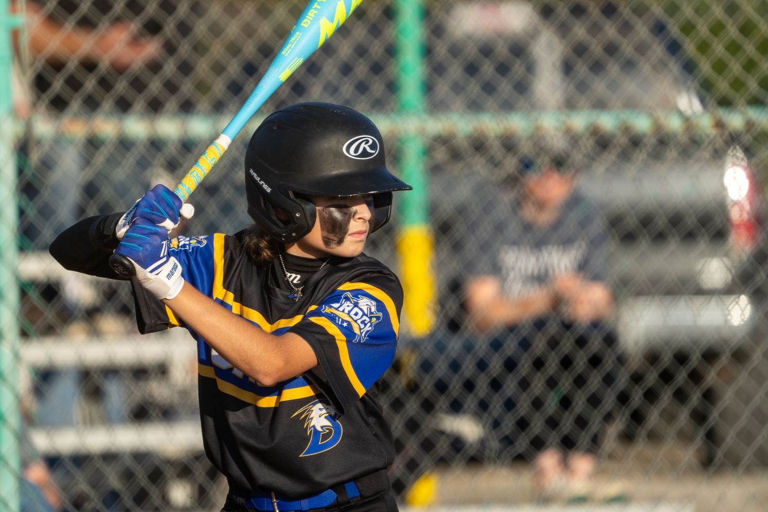 Baseball player wearing a blue and yellow uniform swings a bat during a game through chain link fence.