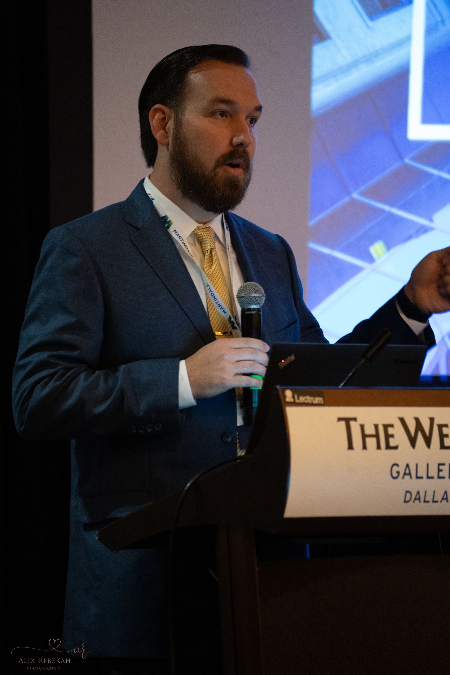 Conference presenter speaking at The Westin Galleria podium with projection screen in background.