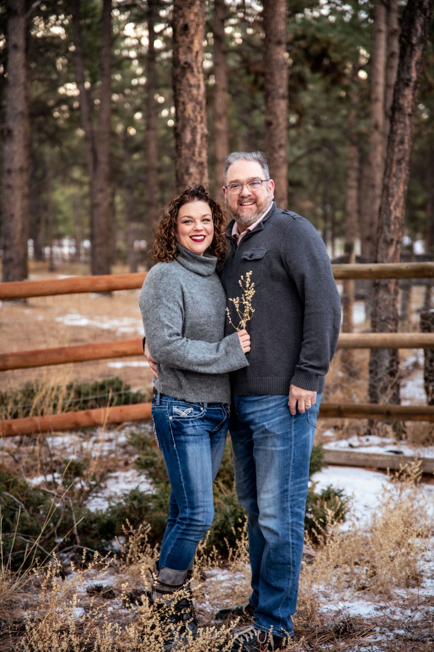 Couple in warm sweaters embracing by wooden fence in snowy pine forest.