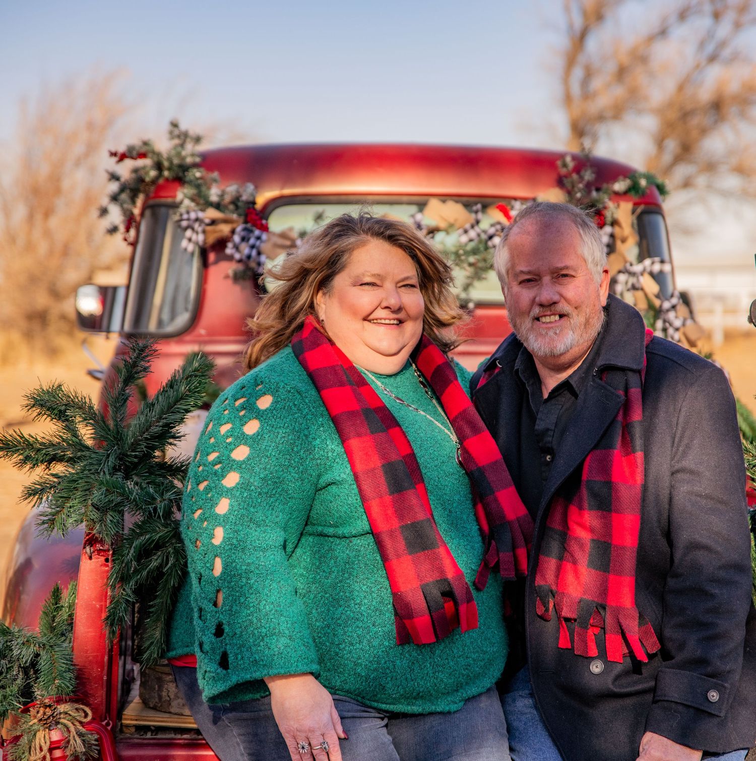 A couple in matching buffalo plaid scarves pose by a vintage red truck decorated with holiday greenery at sunset.