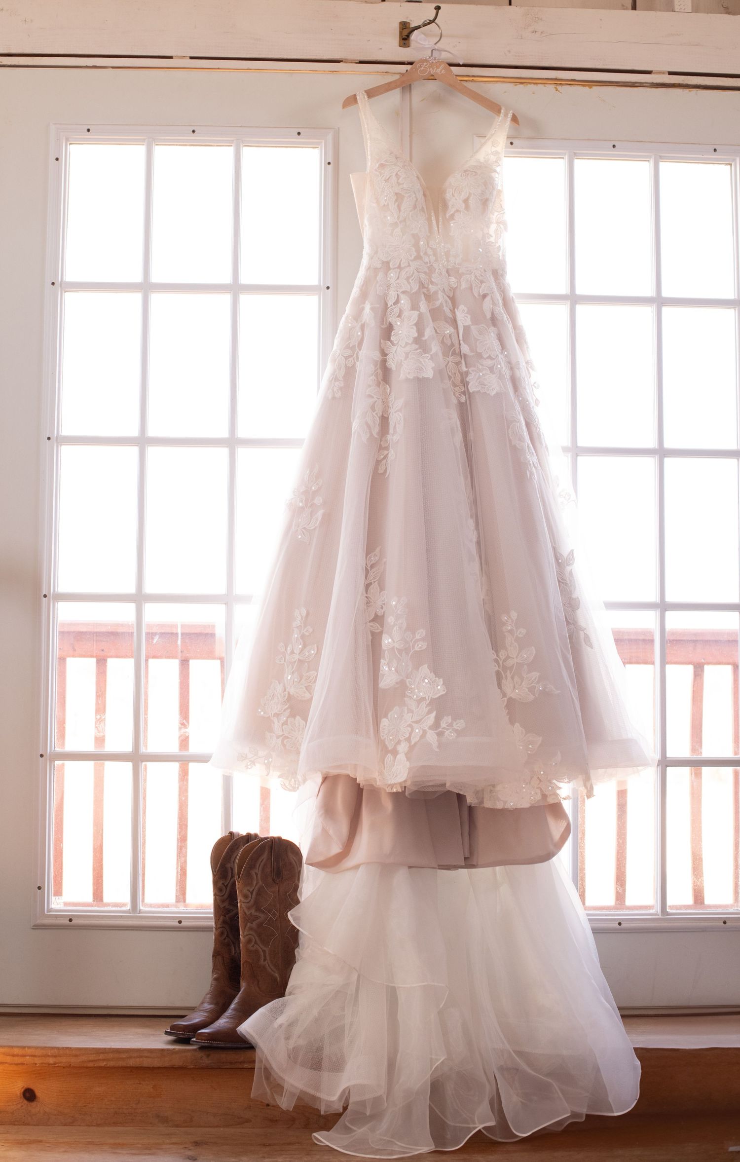 A white lace wedding dress hangs by a window with cowboy boots positioned underneath on hardwood floors.