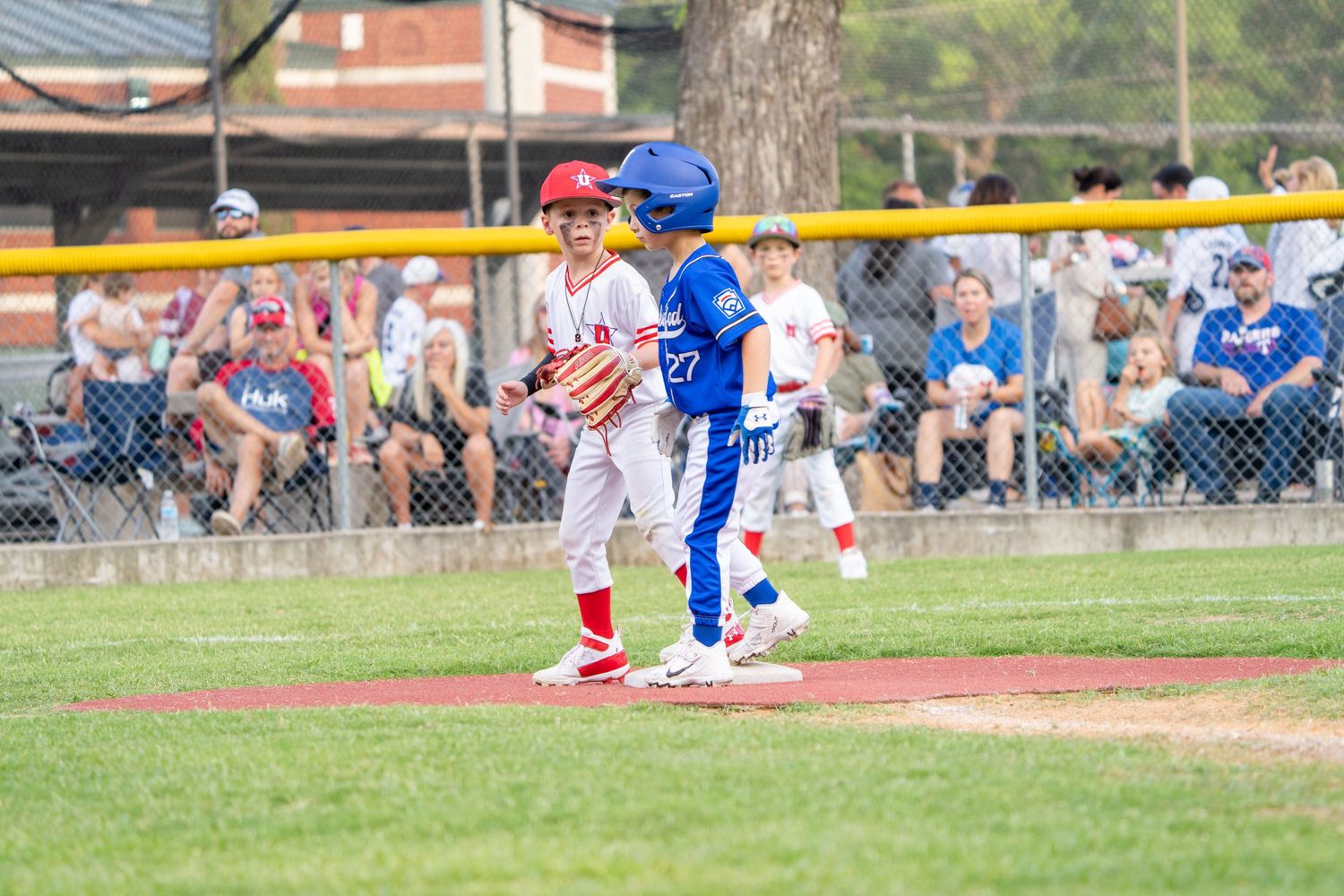 Baseball players in white and blue uniforms compete during a game at an outdoor baseball field on a sunny day.