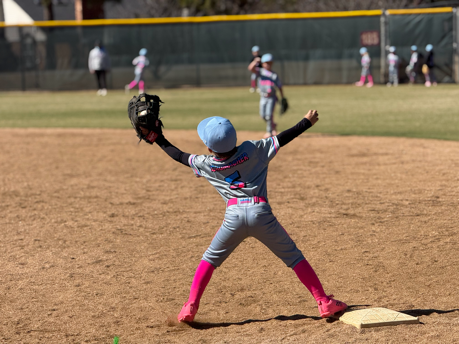 Young baseball player in blue cap and pink socks throwing ball on dirt infield during youth league game.