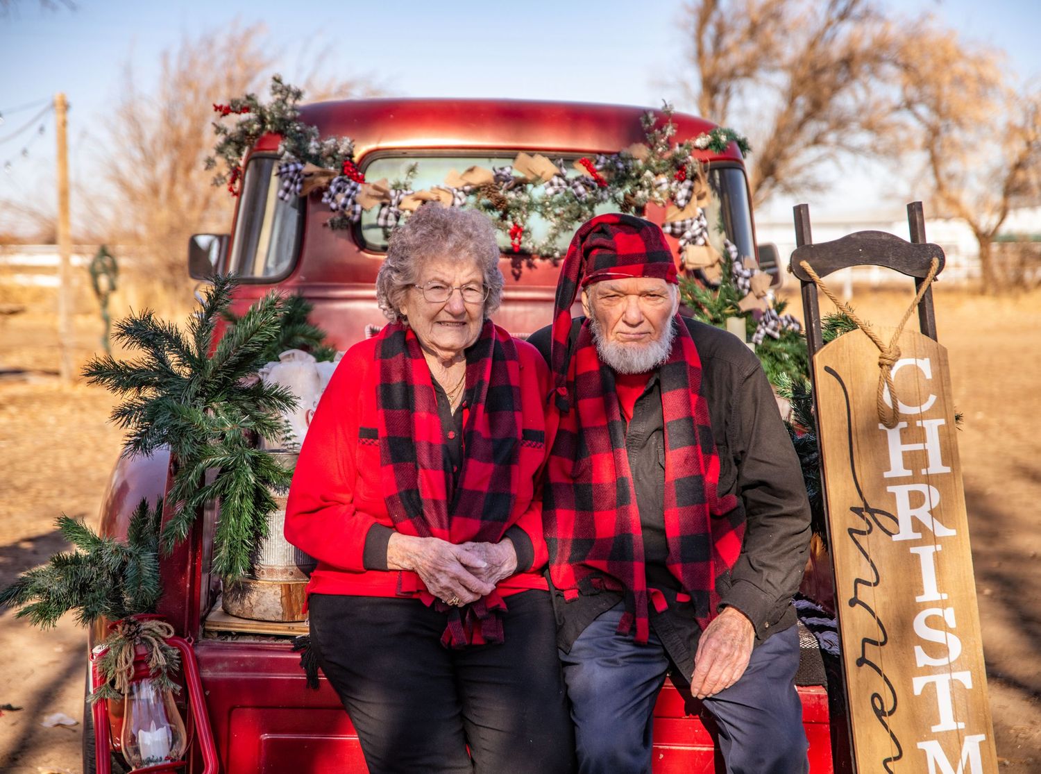 Two people in matching red and black buffalo plaid sitting in a vintage red truck decorated for Christmas.