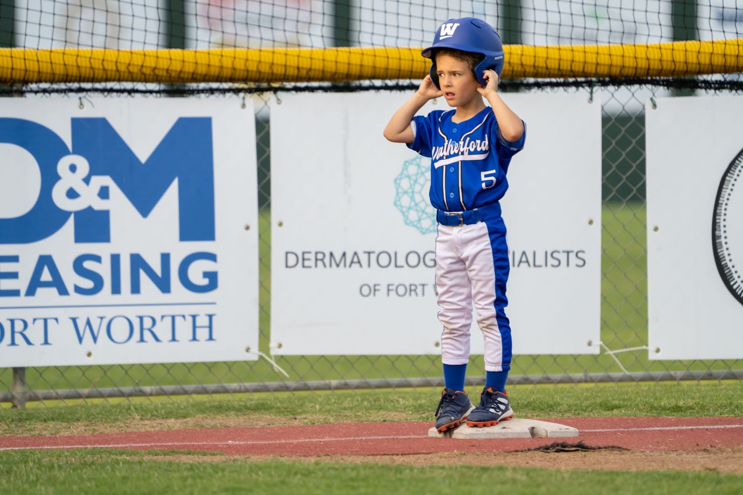 Young baseball player in blue uniform stands on base during youth league game with advertising banners in background.