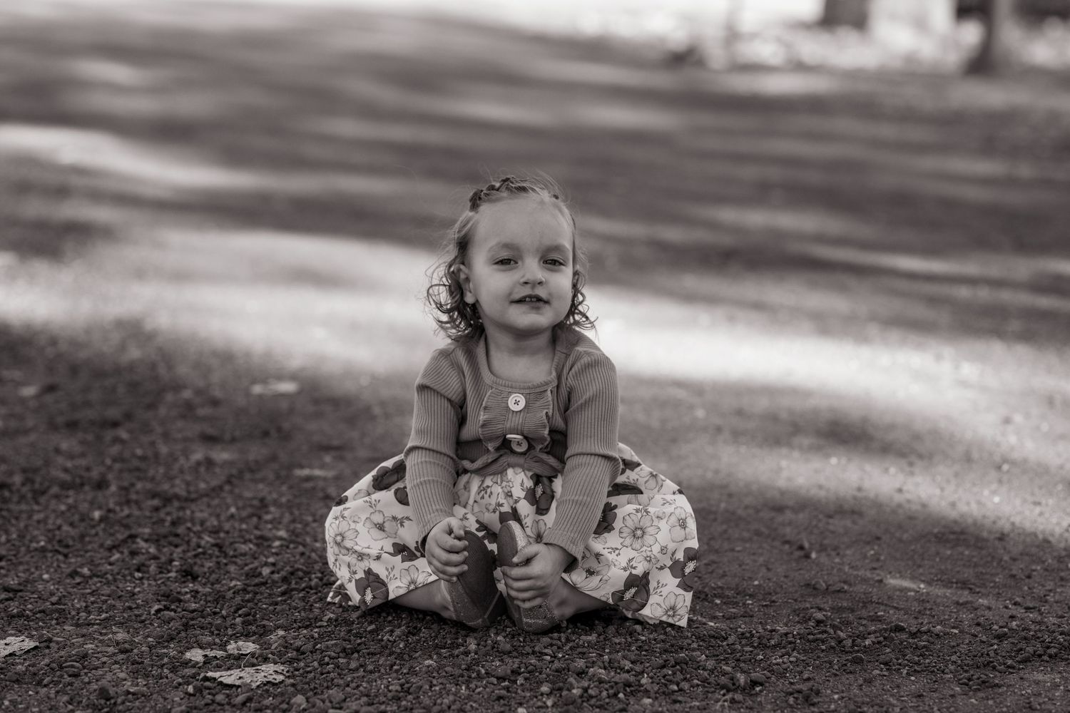 Black and white portrait of a young child sitting on ground wearing floral dress.