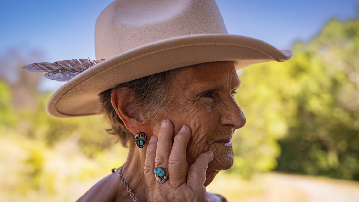 Person in a western hat with turquoise jewelry smiles while touching their face in outdoor lighting.