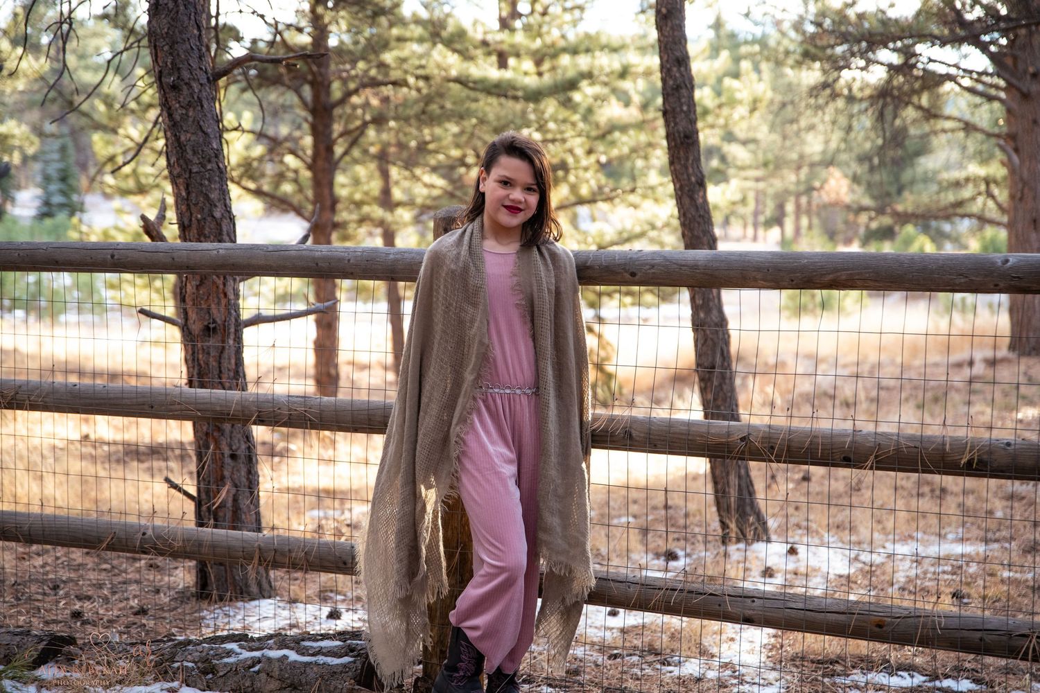 Person in long cardigan and pink outfit standing by wooden fence in winter forest landscape.