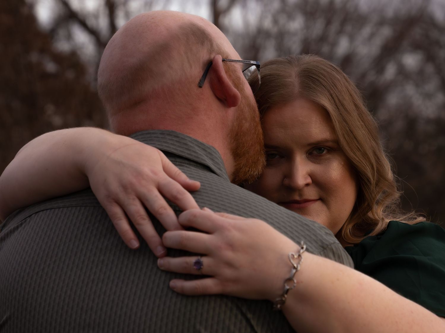A woman smiles warmly while embracing a bald man with a red beard outdoors in a romantic couples portrait.