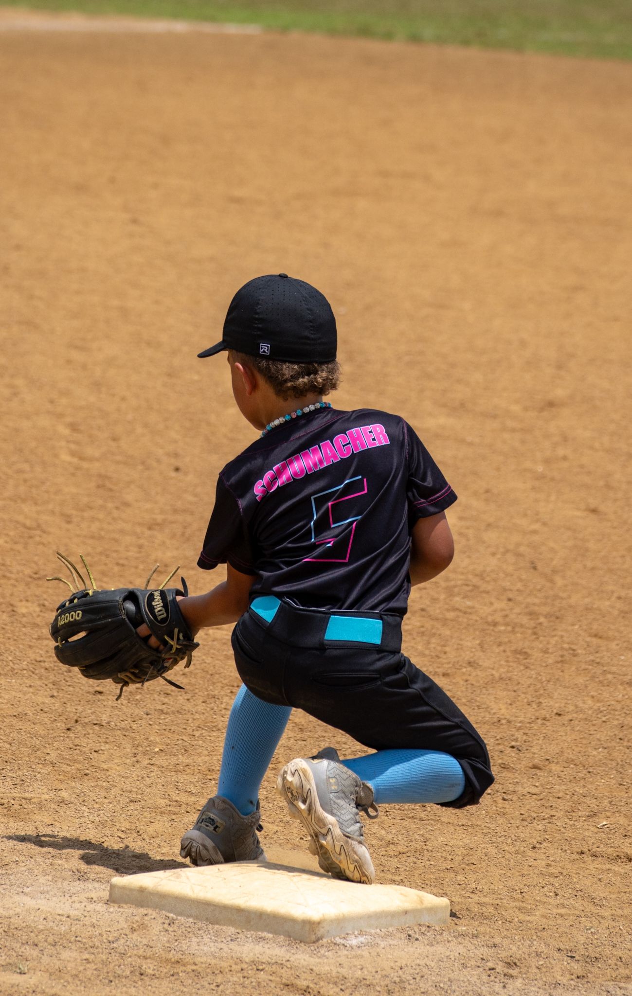 Youth baseball player in black uniform and blue socks kneeling on first base during a game on dirt infield.