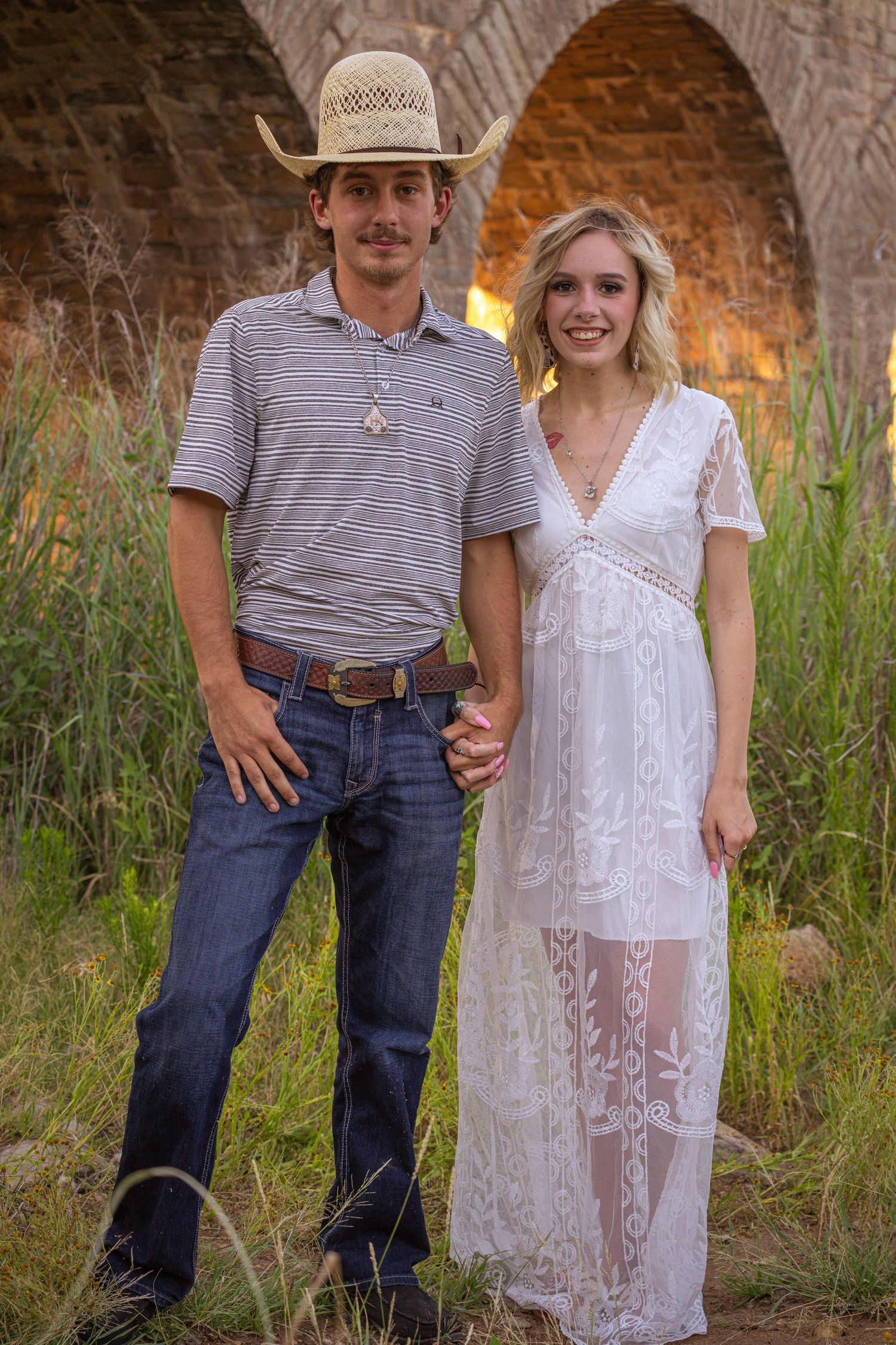 A couple poses together against tall grass at sunset wearing contrasting white dress and casual striped shirt with jeans.
