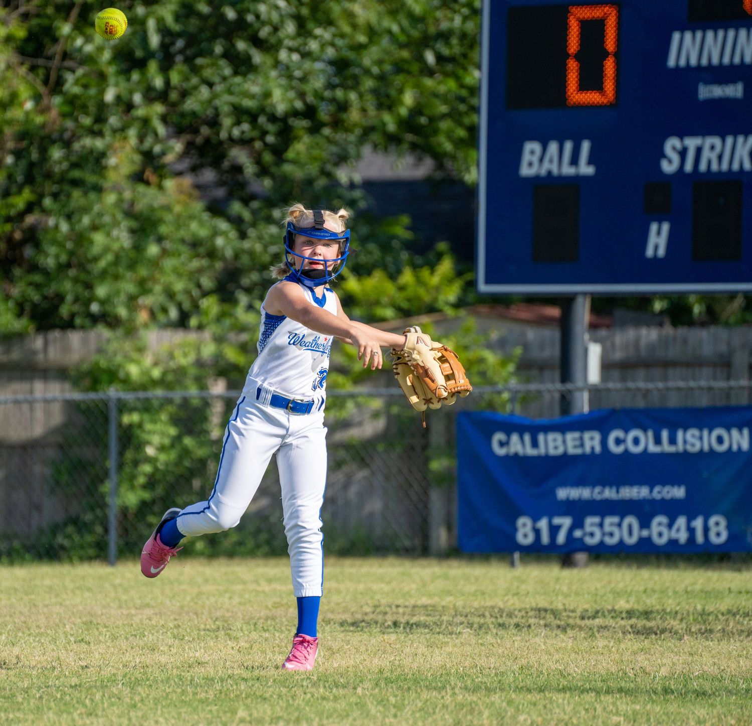 A softball player in a blue and white uniform throws a yellow ball in a grassy outfield with a scoreboard visible.