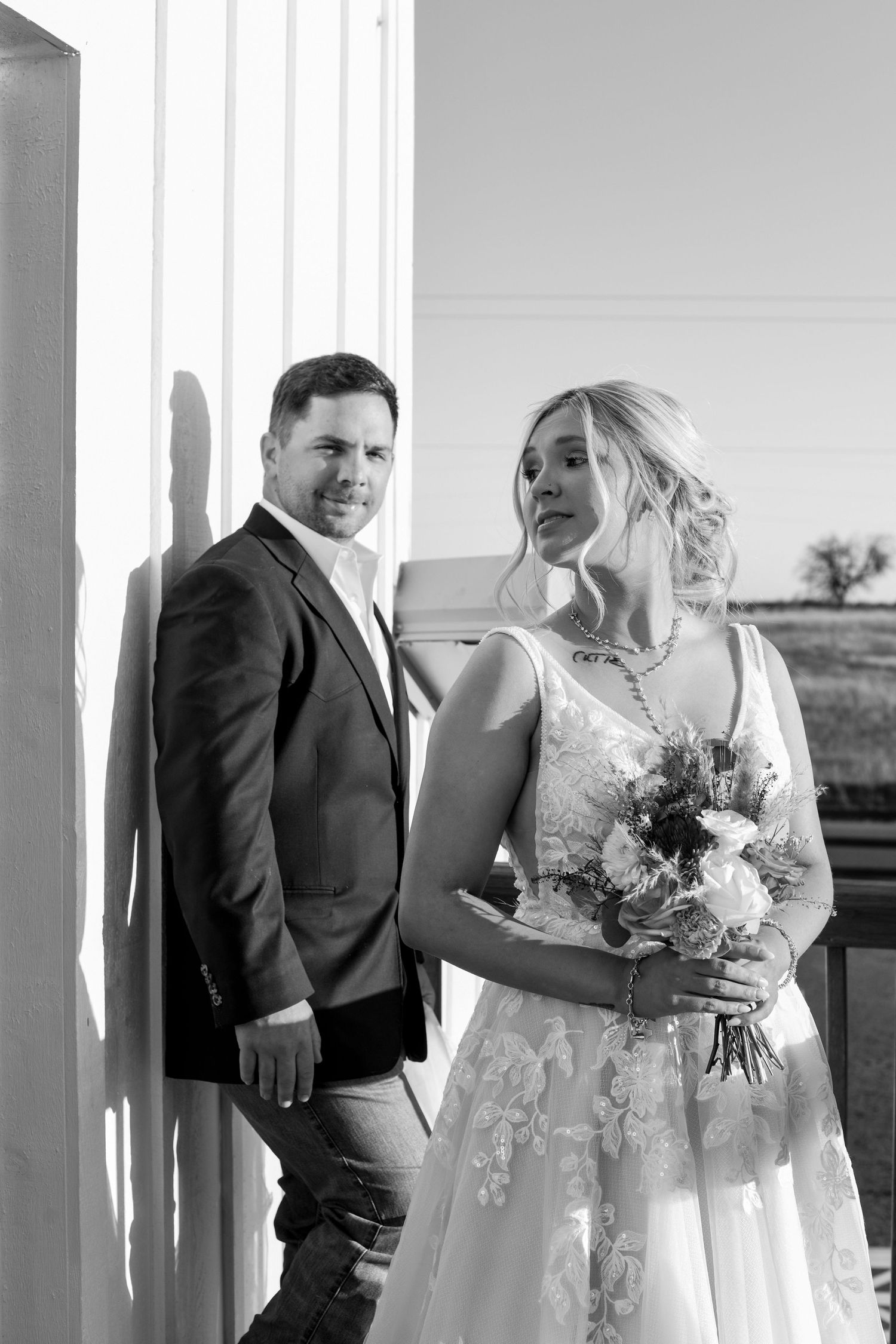 A black and white wedding portrait captures a couple in formal attire with a bridal bouquet against a light wall.