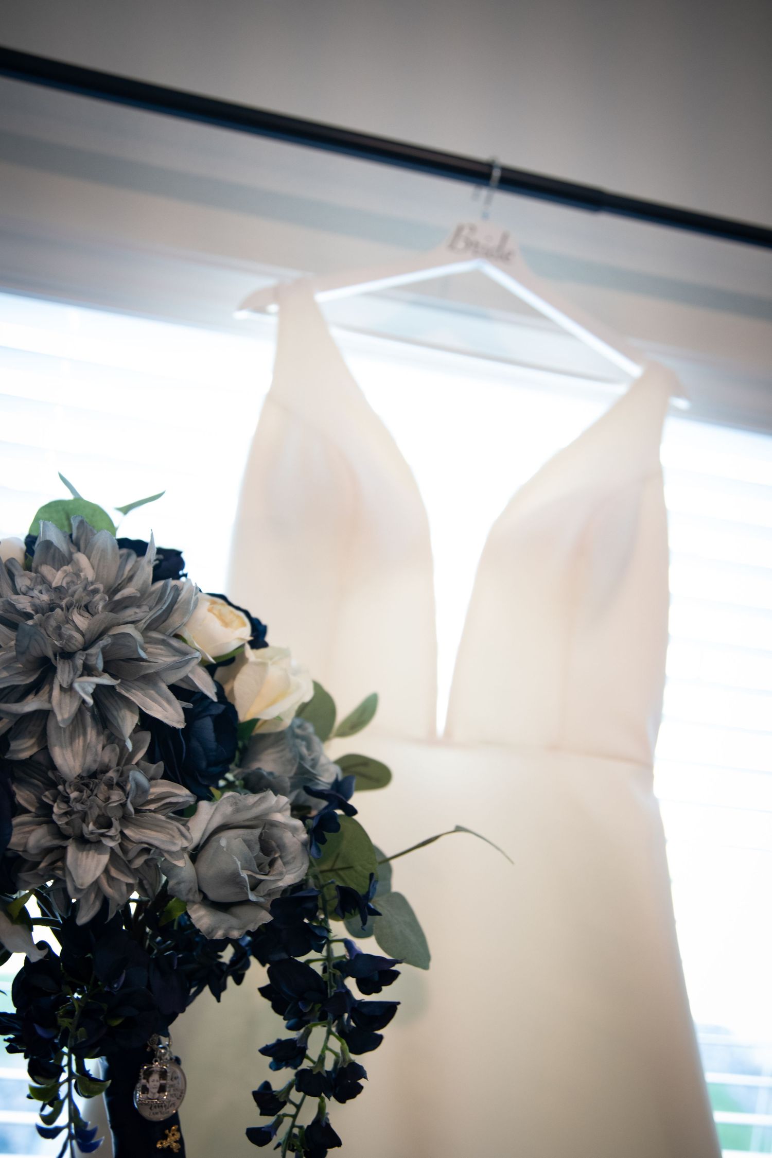 White wedding dress on hanger with gray floral bouquet against window.