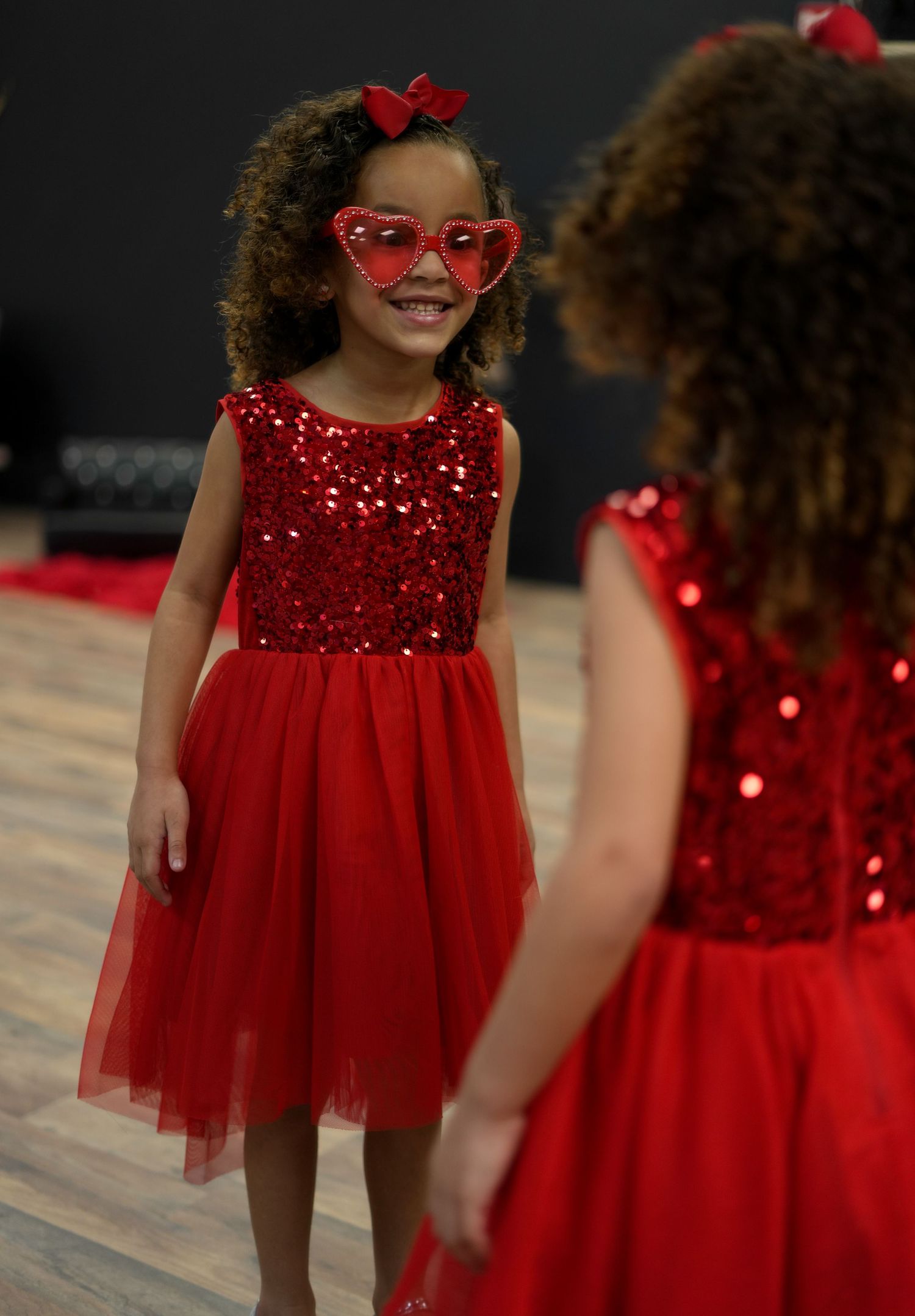 Child in sparkly red dress with heart-shaped sunglasses and bow smiles while standing in a festive setting.