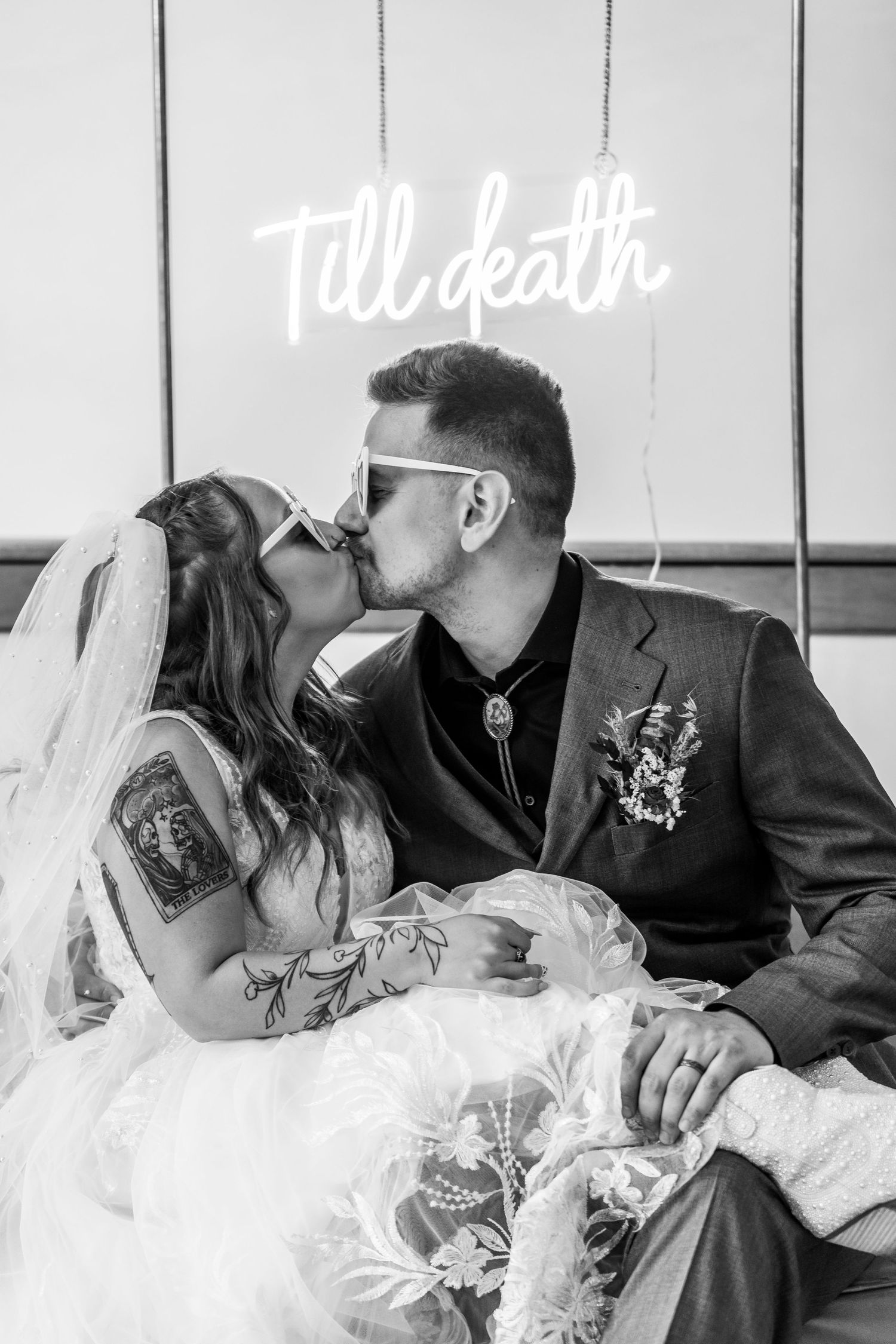 Romantic moment under neon 'Till death' sign in black and white photography.