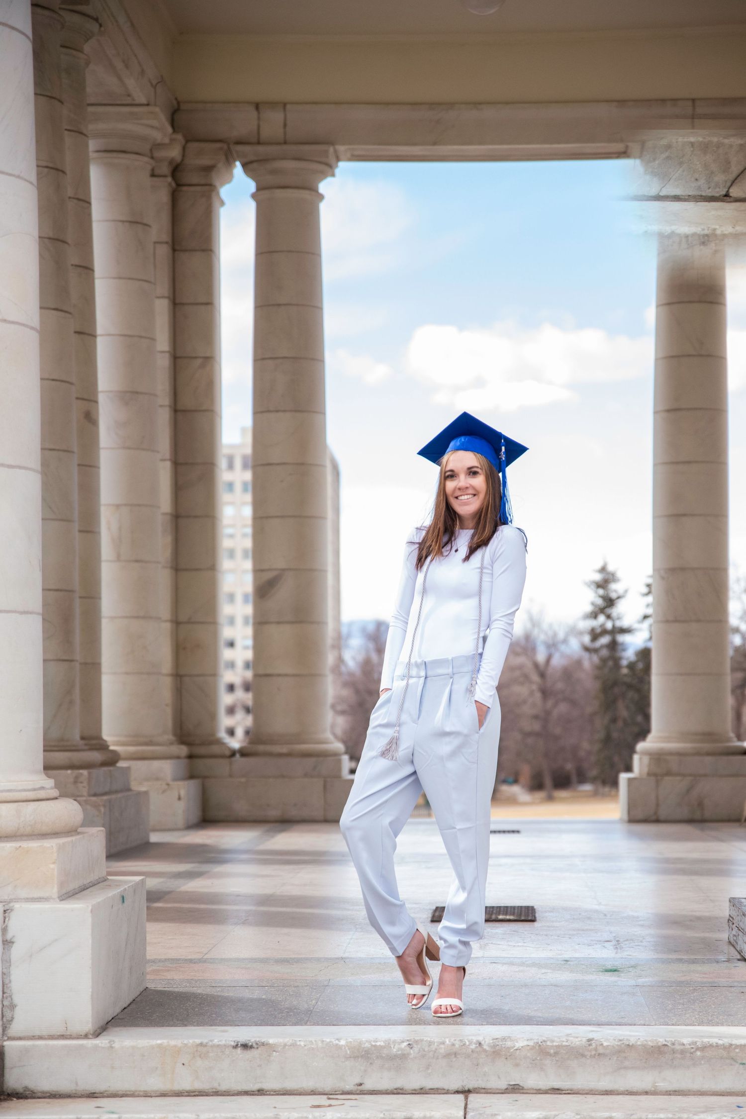 Graduate in white outfit and blue cap poses elegantly between ornate columns.