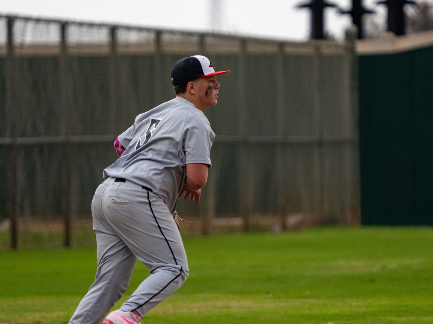 Baseball player in gray uniform and red cap runs across green outfield during a game.