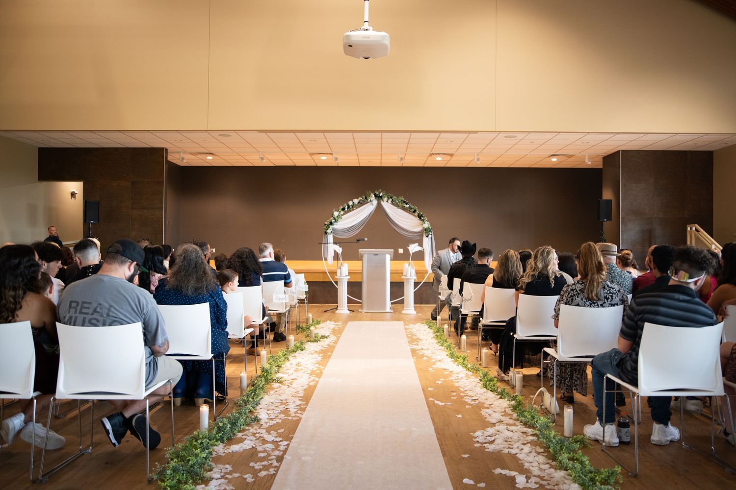 Wedding ceremony setup in hall with white chairs and aisle runner leading to arch.