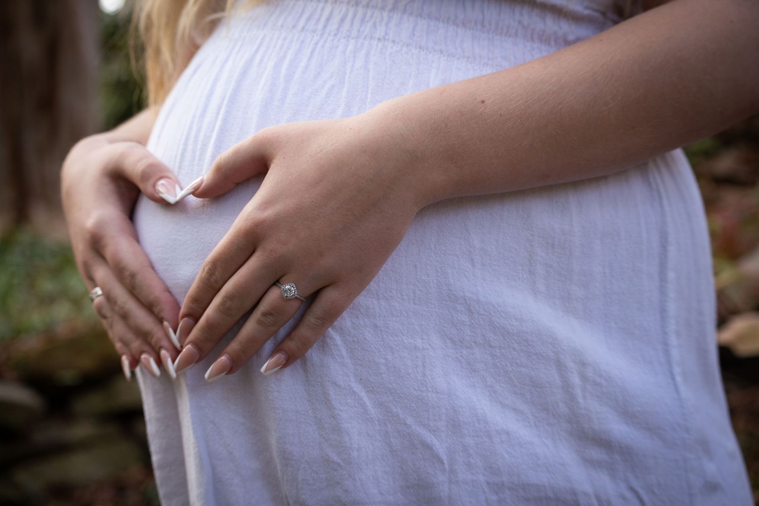 Hands form a heart shape over a pregnant belly wearing a white dress, with nature blurred in the background.