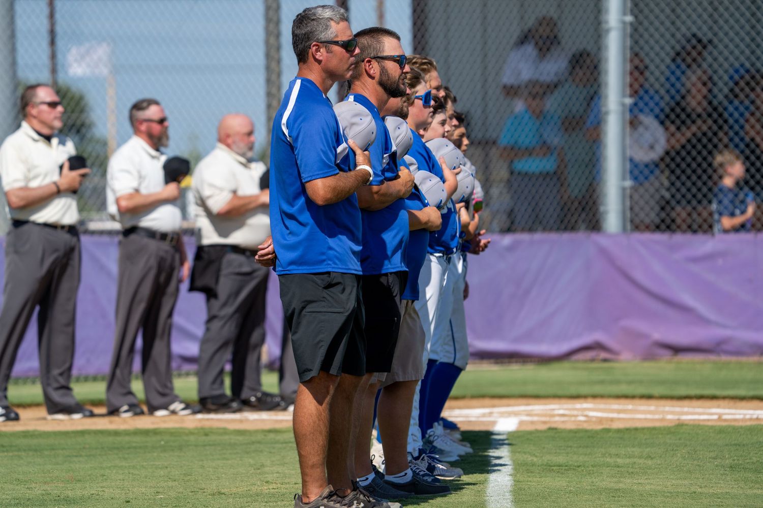 Baseball coaches line up along the field during a game while spectators observe in the background.