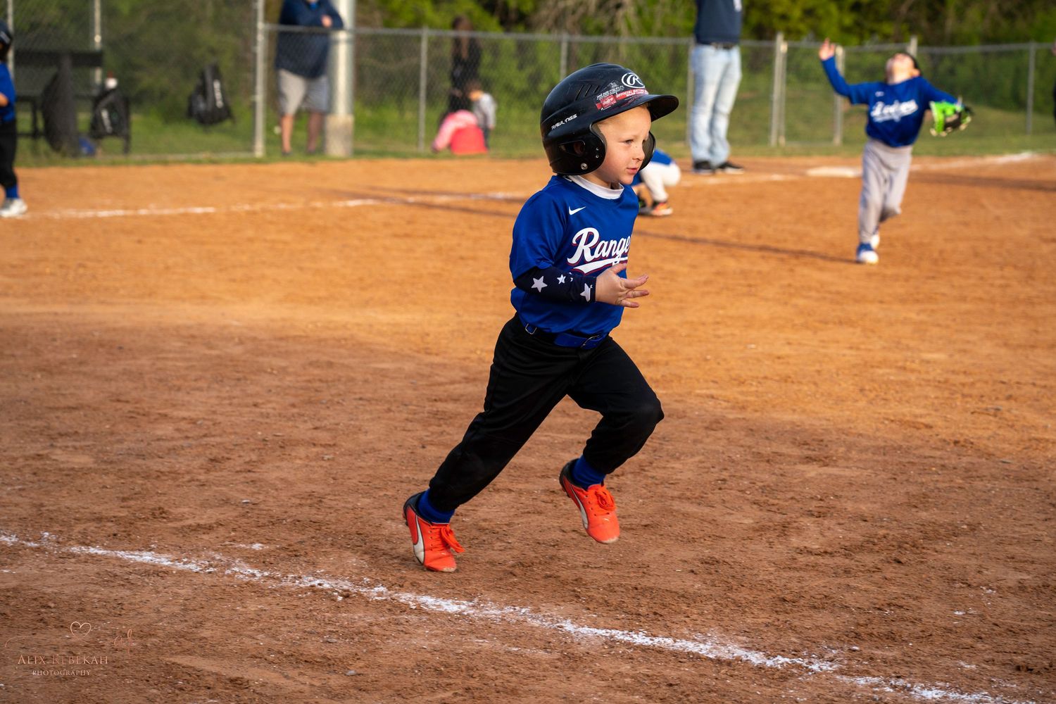 Young baseball player in blue jersey and black pants runs on dirt baseline during youth baseball game.