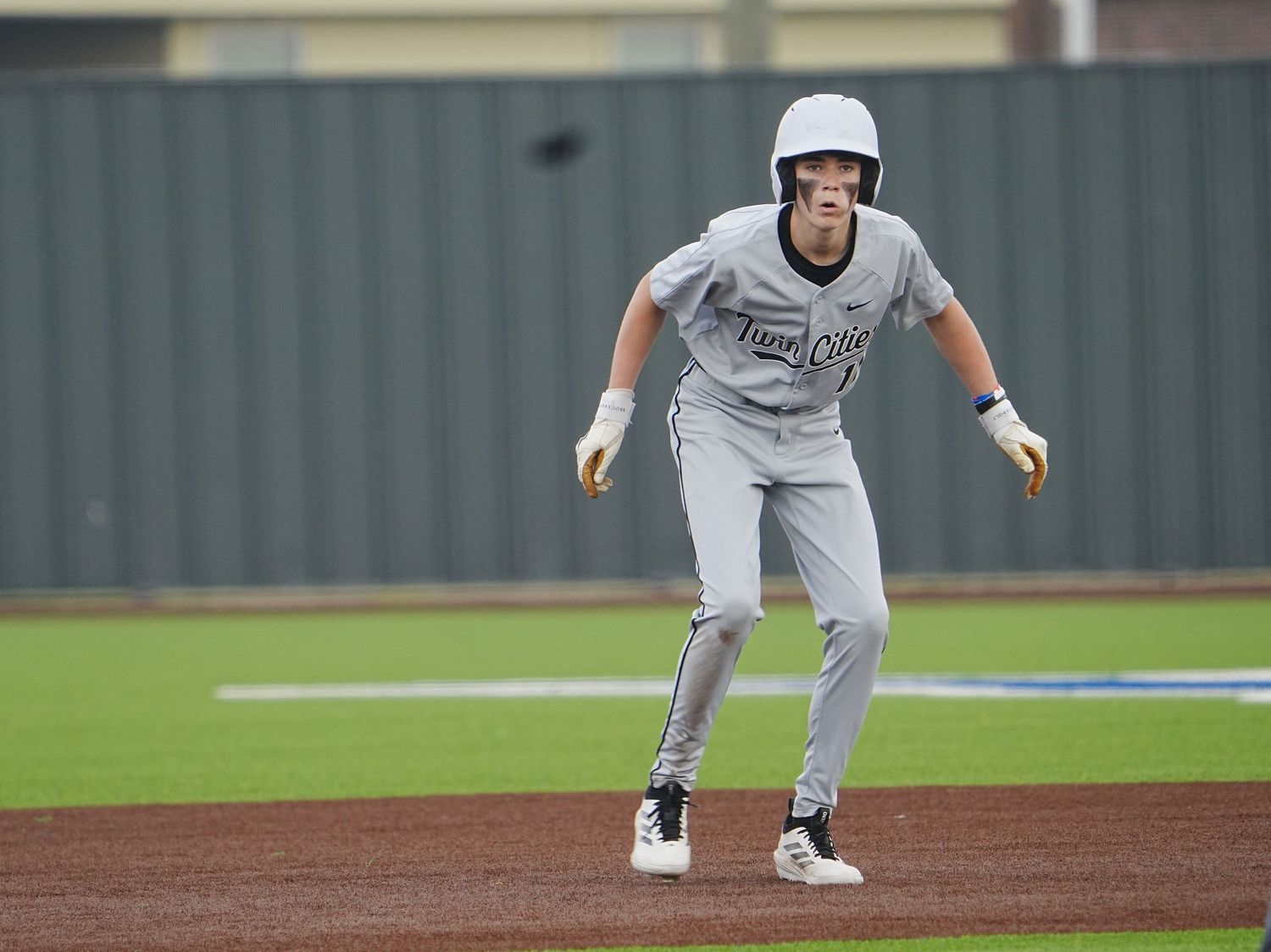 Baseball player in gray uniform leads off base during a game, crouching in ready stance on the field.